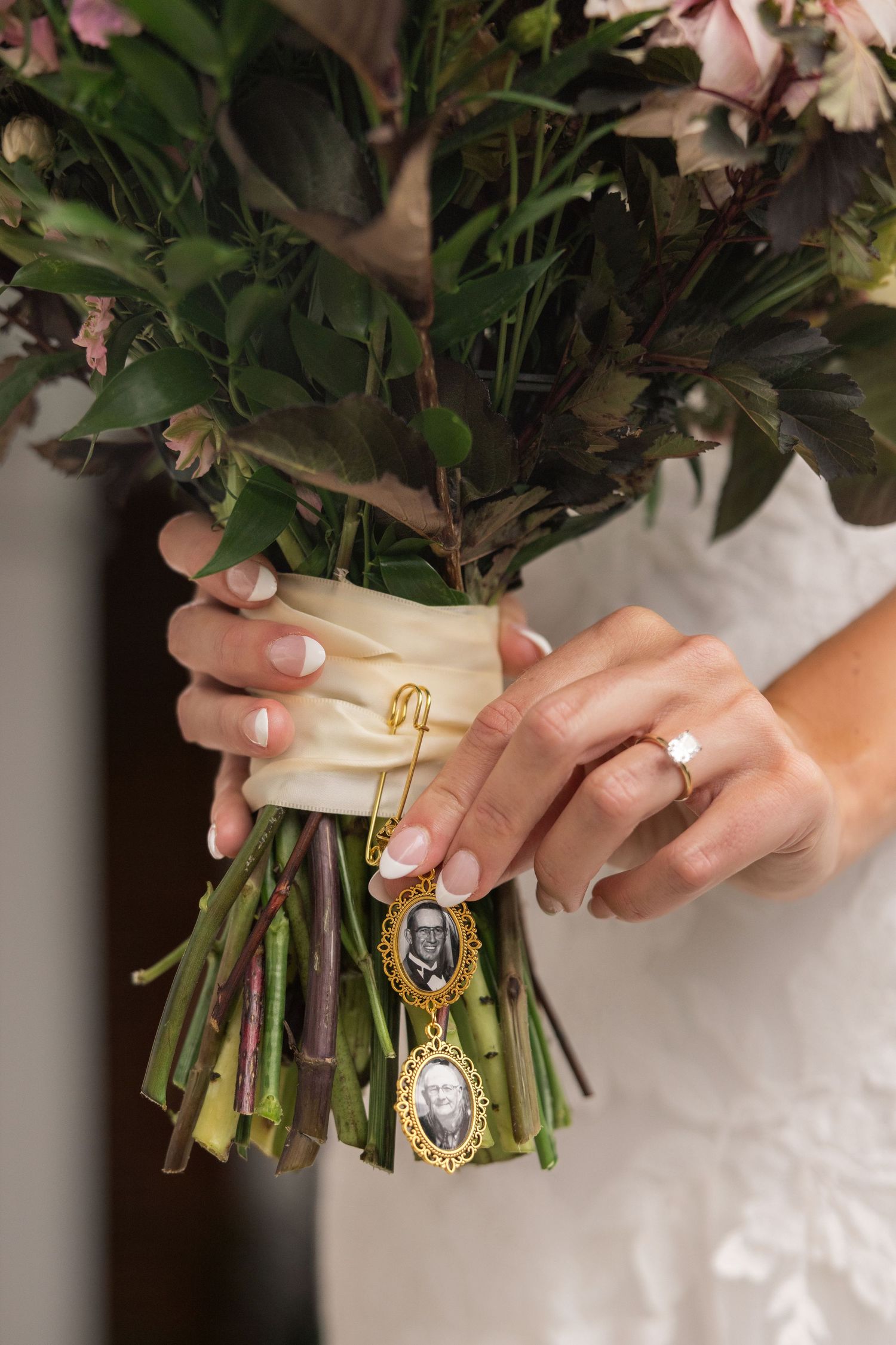 Bride holding floral bouquet with gold memorial photo charms and safety pin, wearing engagement ring and white dress.