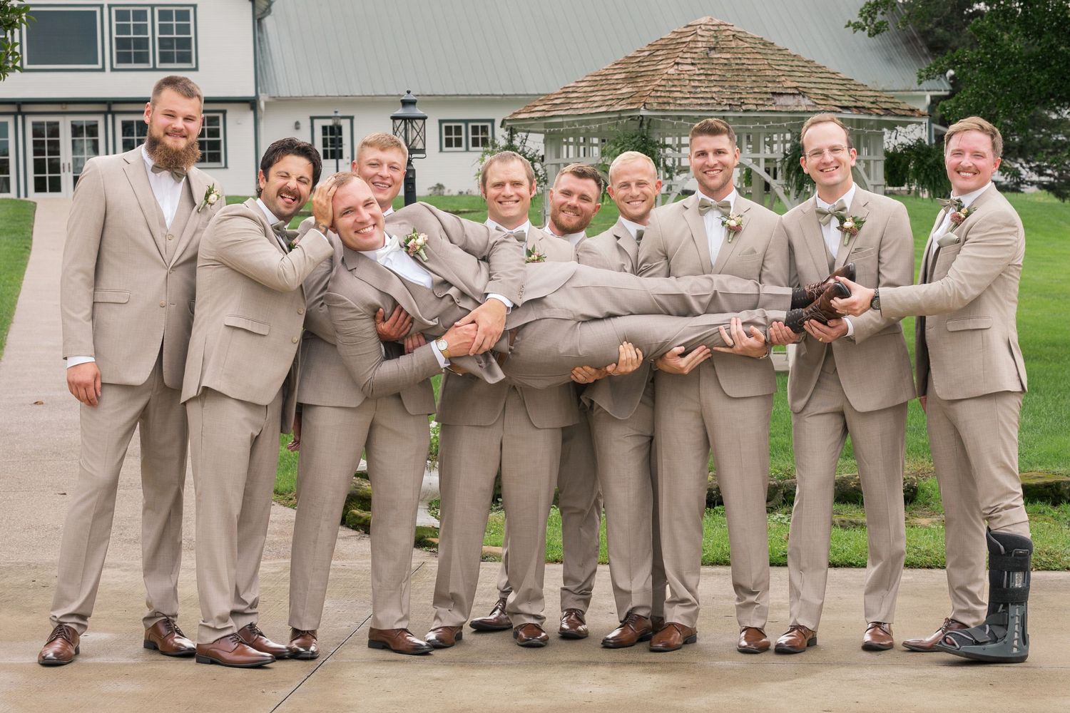 Groomsmen in tan suits lifting the groom in a fun outdoor wedding photo at a white venue.