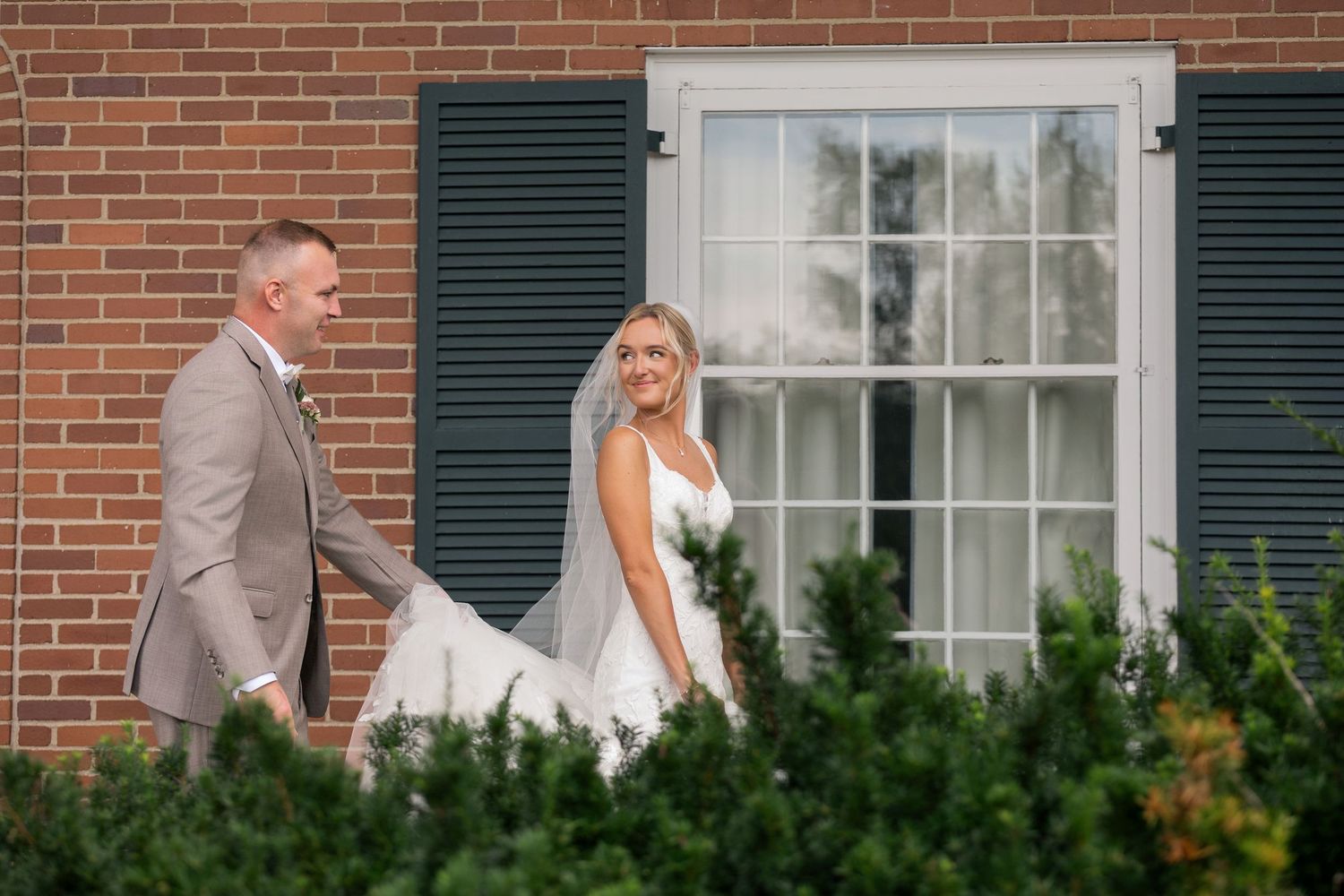 Bride and groom walking together outside a brick building with dark shutters on their wedding day.