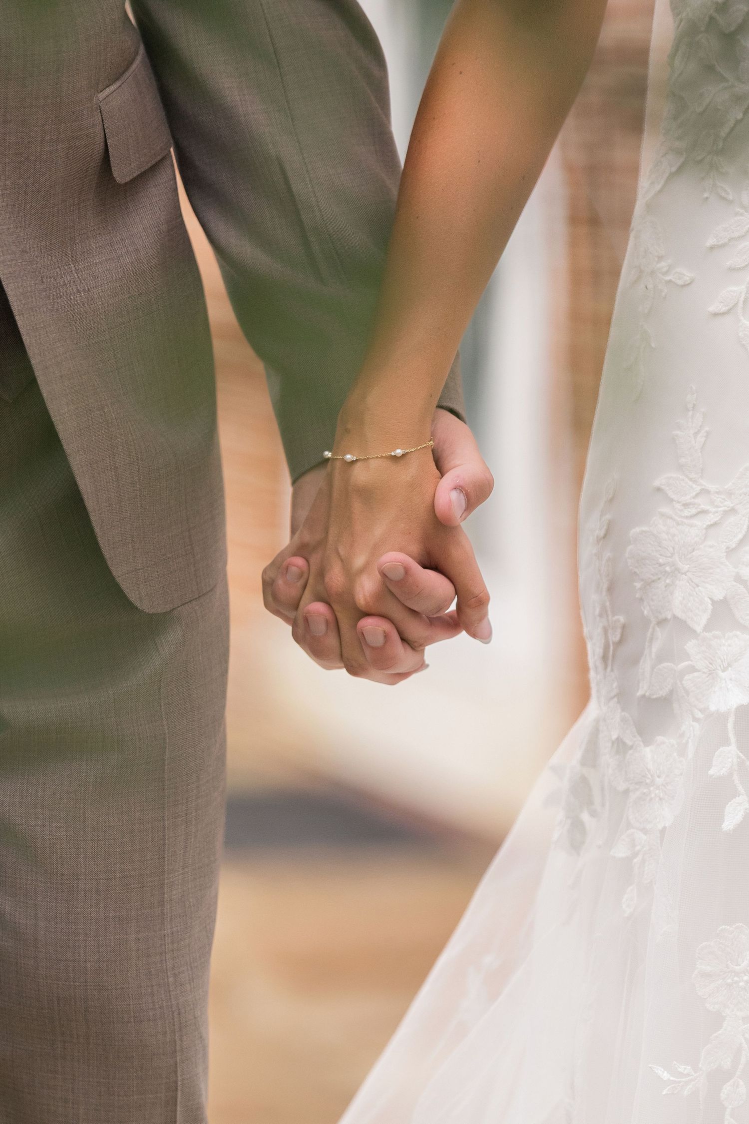 Bride and groom holding hands at wedding, bride in lace dress with bracelet, groom in gray suit.