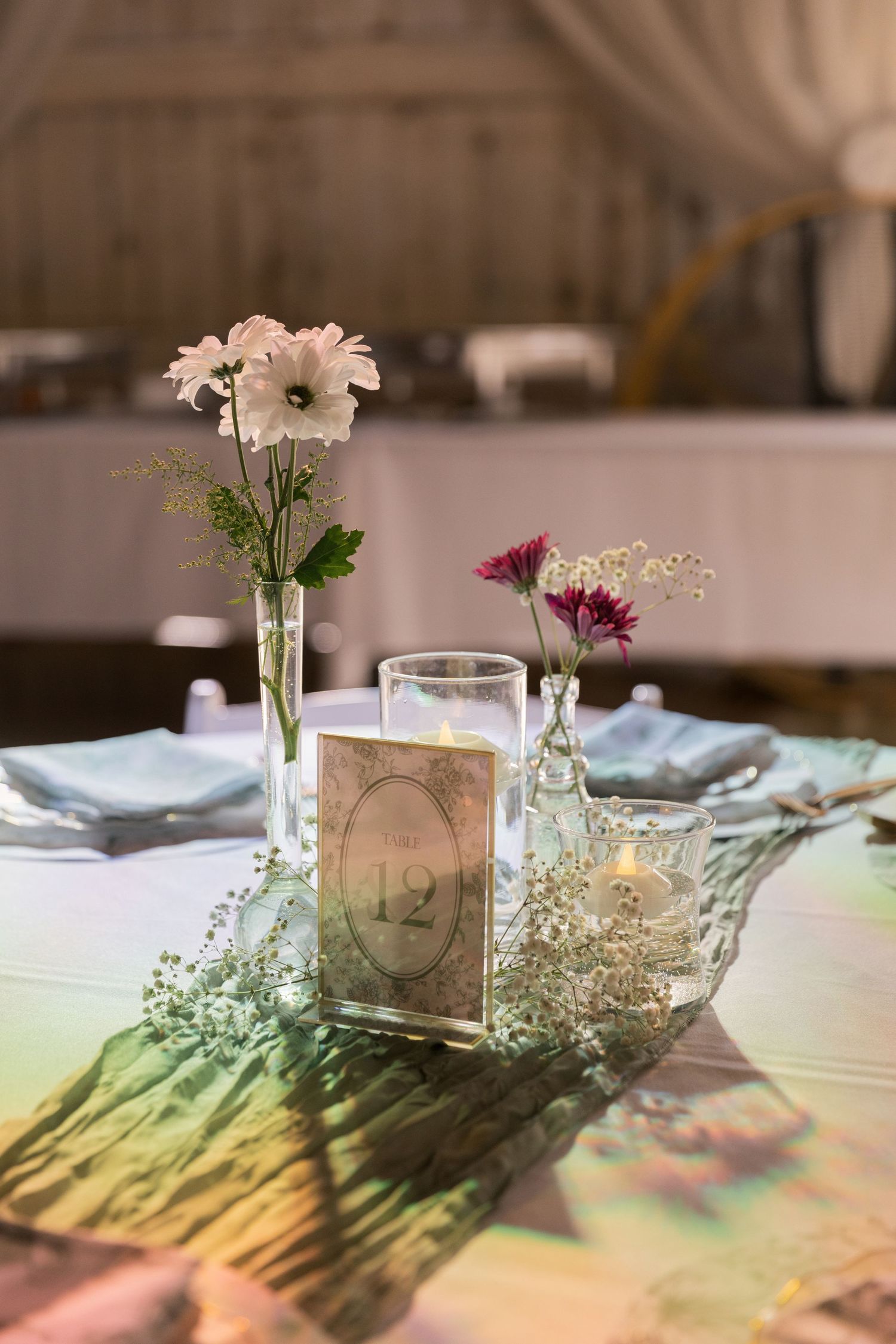 Elegant wedding table centerpiece with white and purple flowers in glass vases, candles, and a number 12 card on white linen.