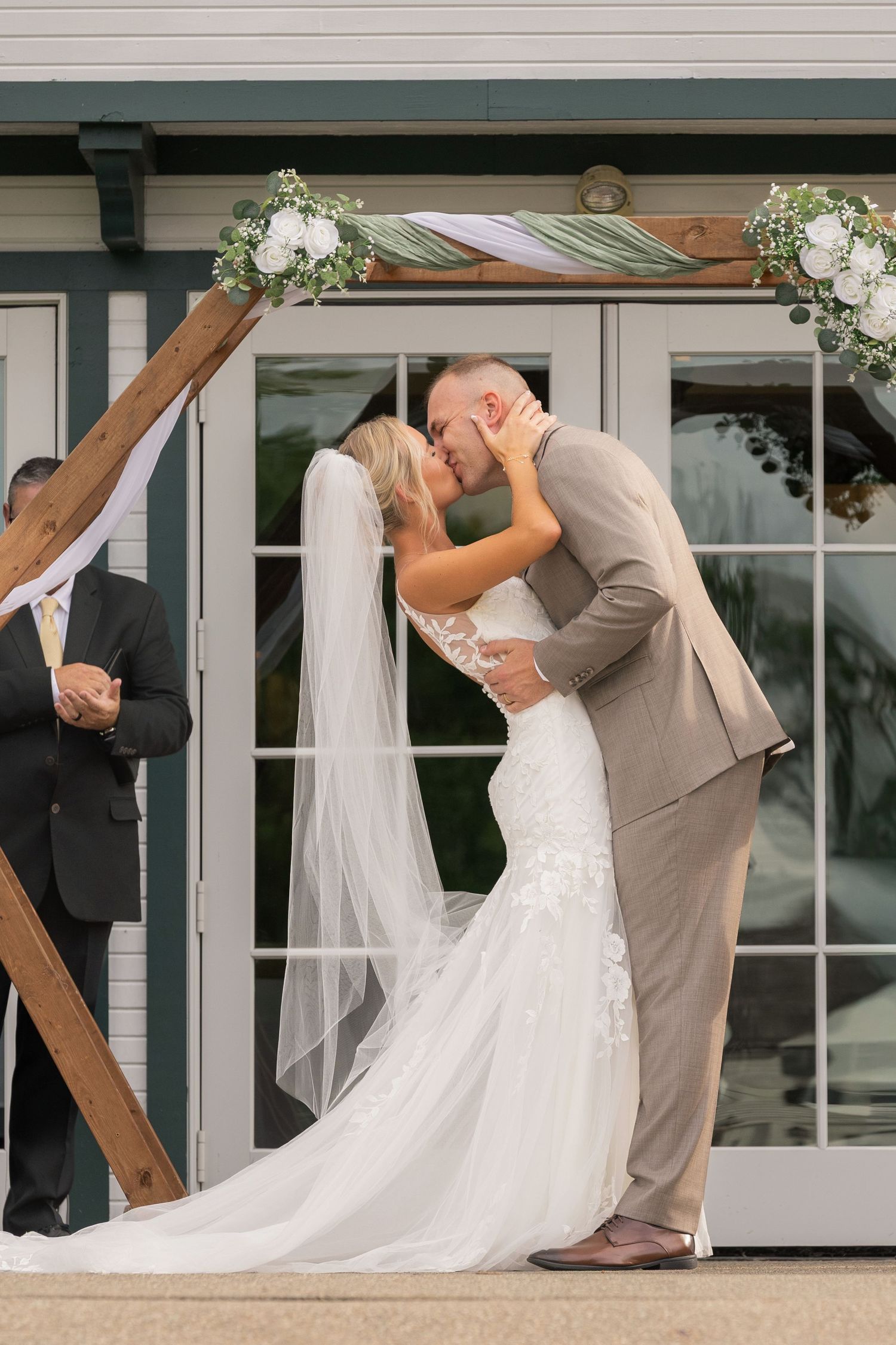 Bride and groom share first kiss under rustic wooden arch with white floral decor at outdoor wedding ceremony.