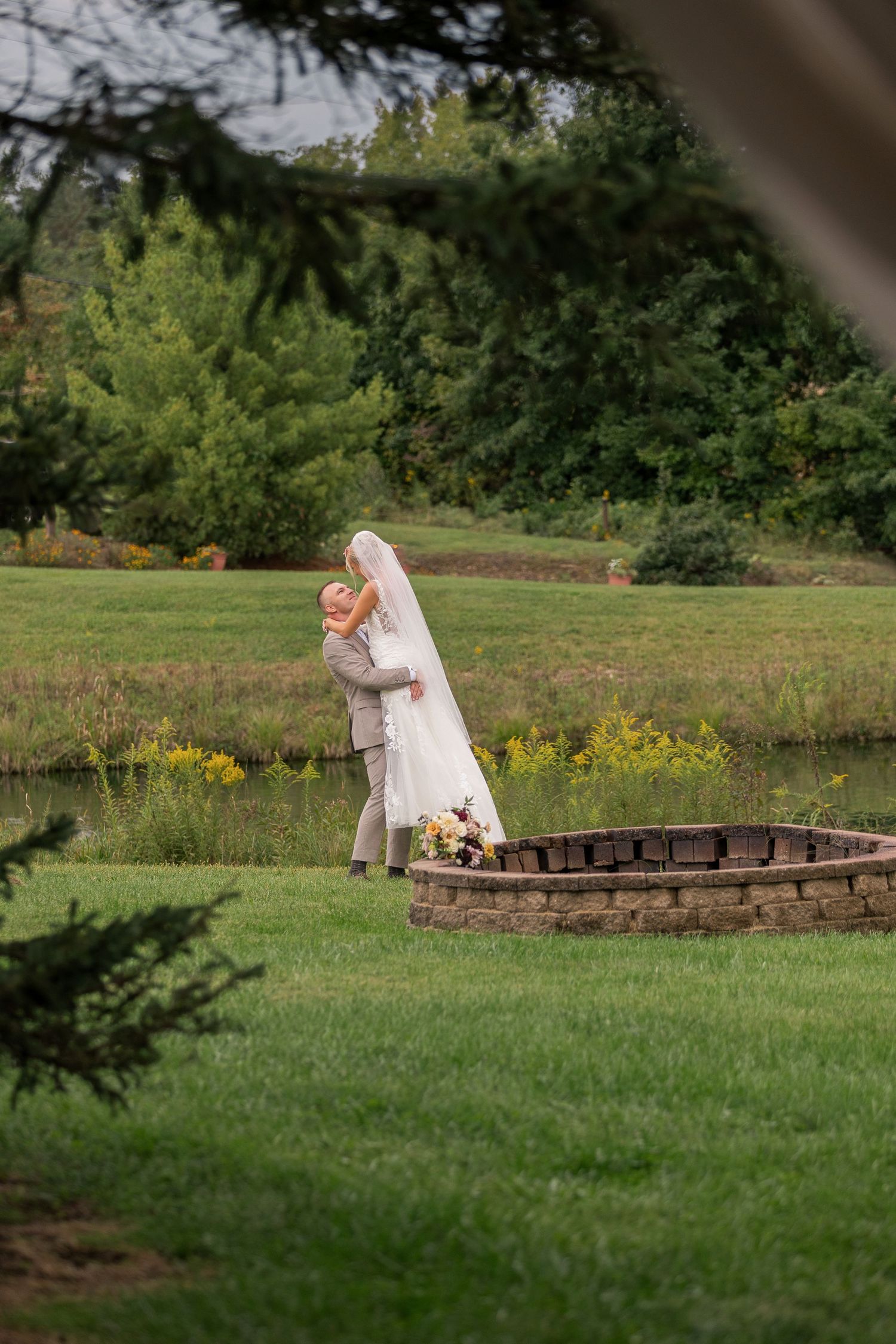 Bride and groom embrace near a stone fire pit in a lush green garden with a pond backdrop on their wedding day.
