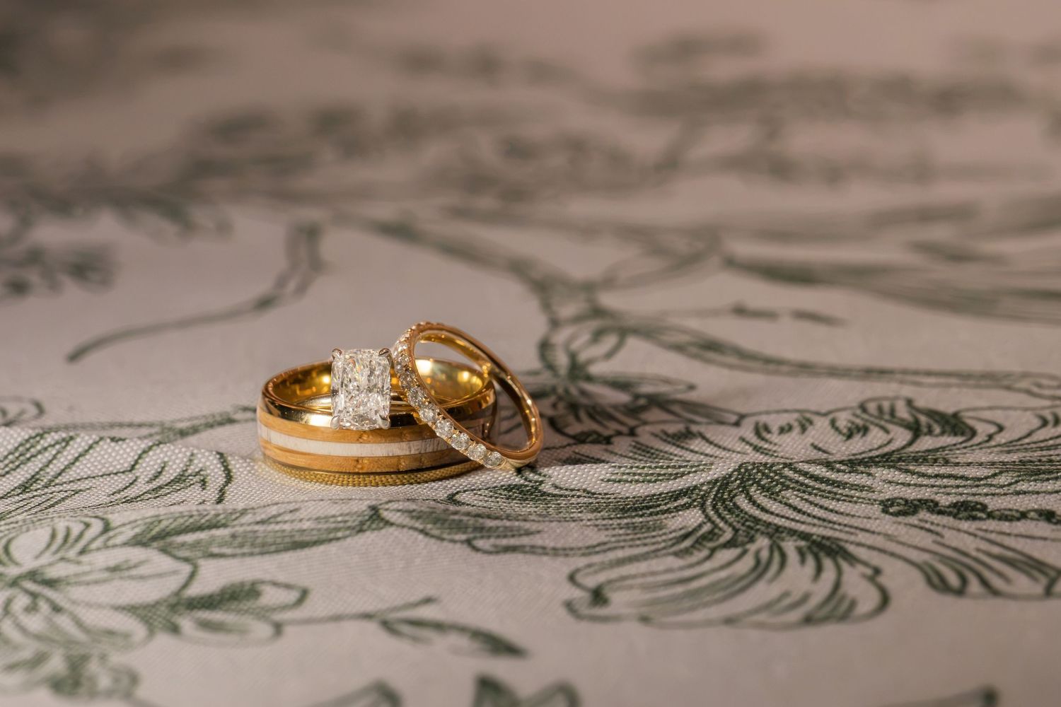 Two gold wedding rings and a diamond engagement ring resting on an elegant floral patterned surface.