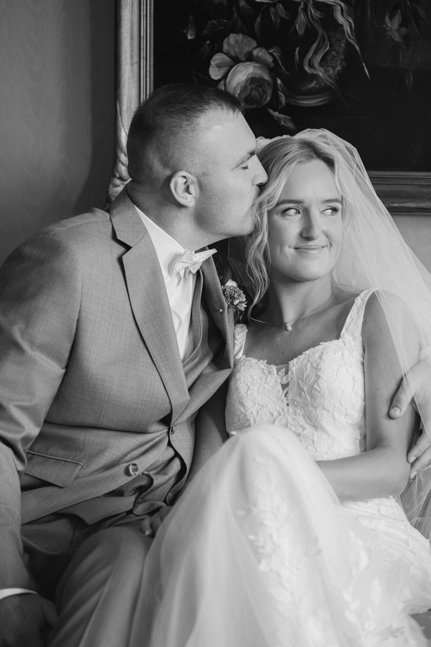 Black and white wedding photo of groom kissing bride on cheek, bride in lace gown and veil smiling softly.