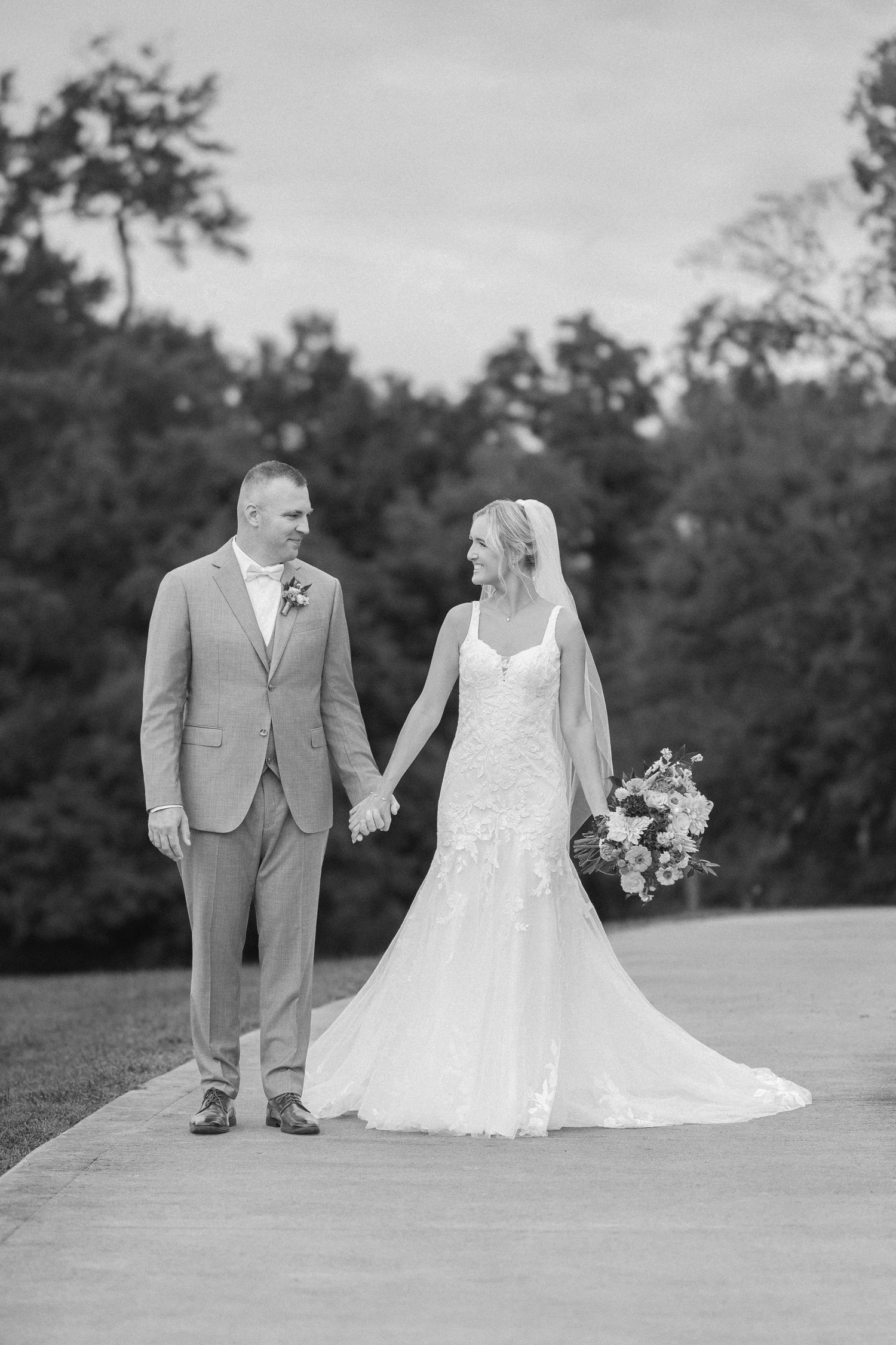 Black and white photo of a bride and groom holding hands on a path, surrounded by lush trees on their wedding day.