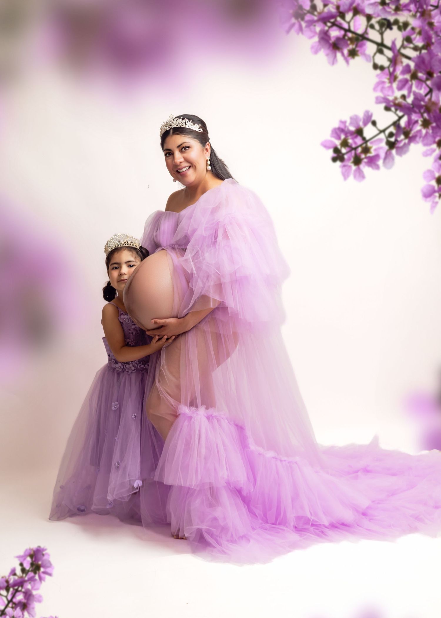 Pregnant woman in hot pink ruffle gown with tiara poses before floral backdrop in elegant maternity photo.