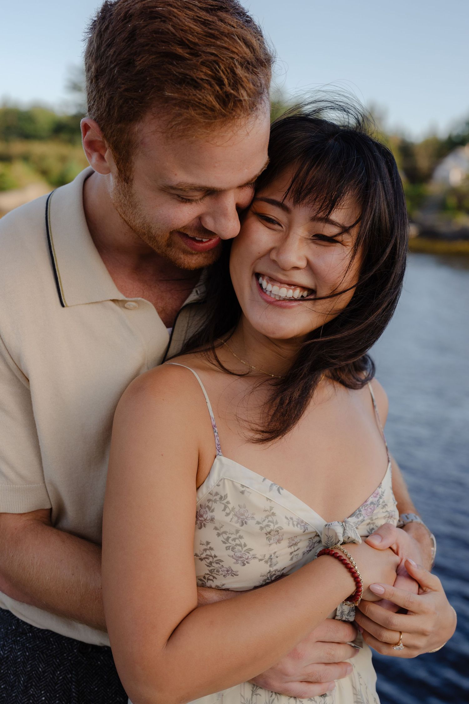 Happy couple embracing outdoors near a lake, woman laughing joyfully in floral dress, man hugging her from behind.