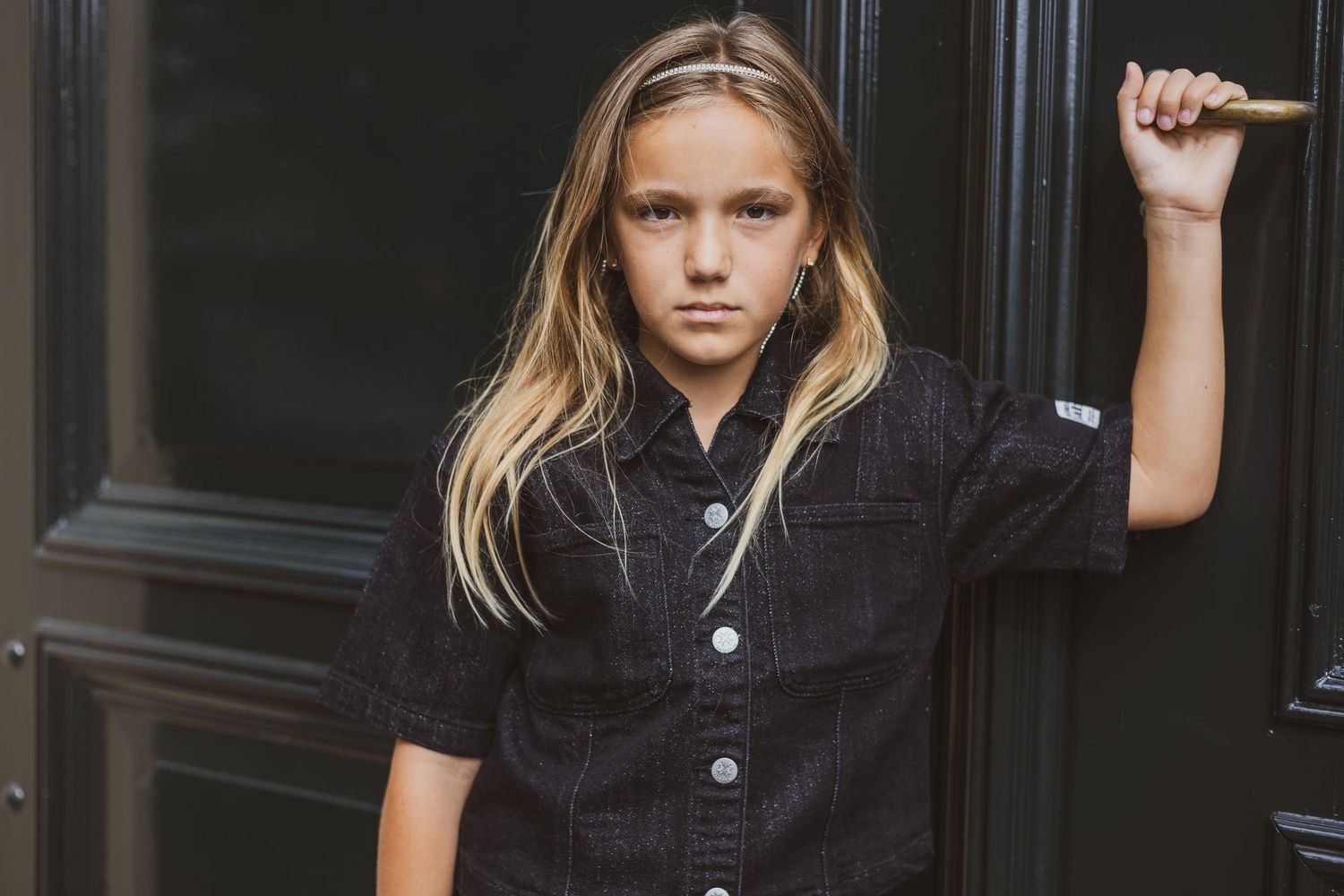 Child model in black denim shirt posing confidently against dark door, long blonde hair, casual fashion shoot.