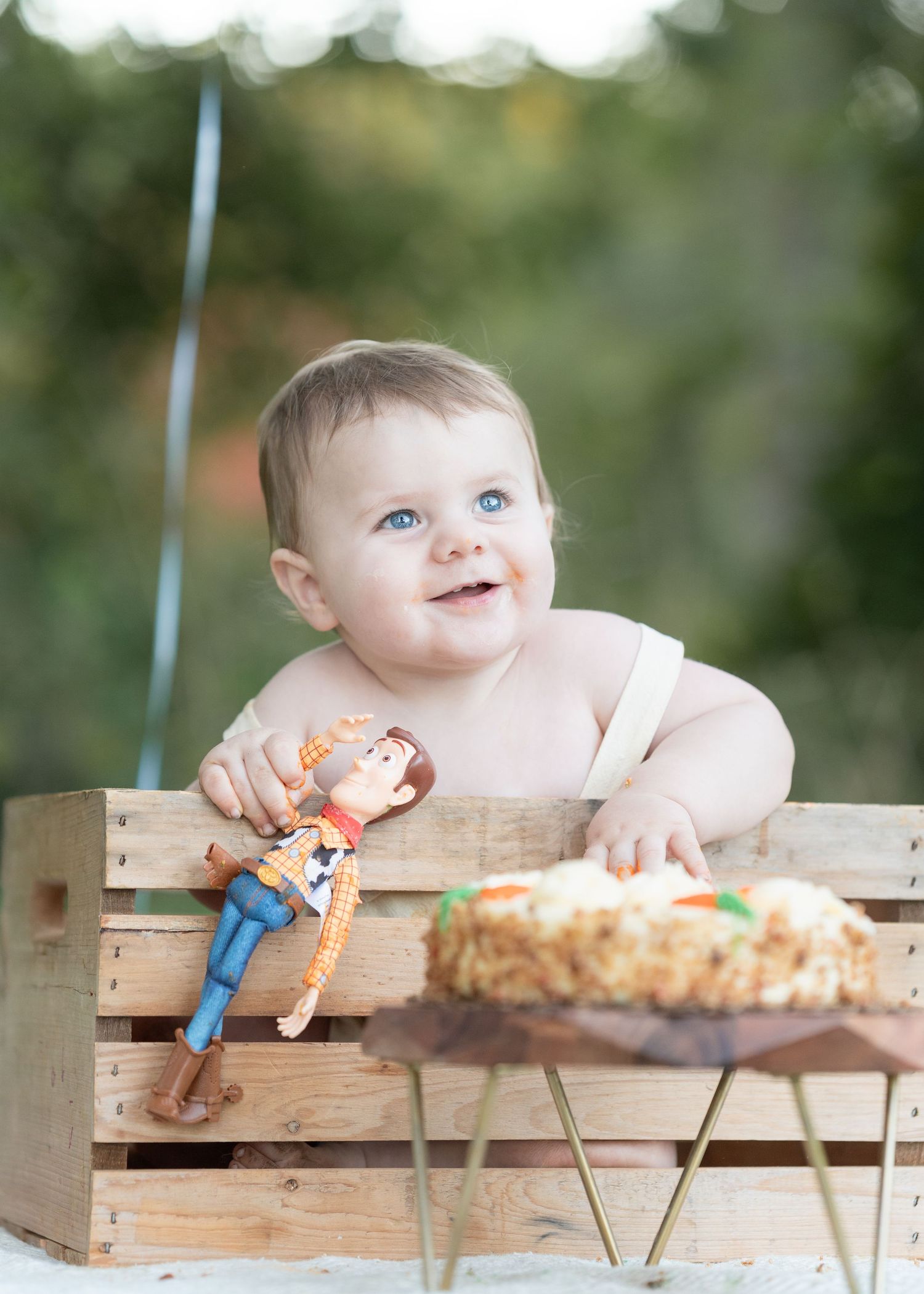 Blue-eyed baby smiling in wooden crate holding Woody toy beside smash cake outdoors.
