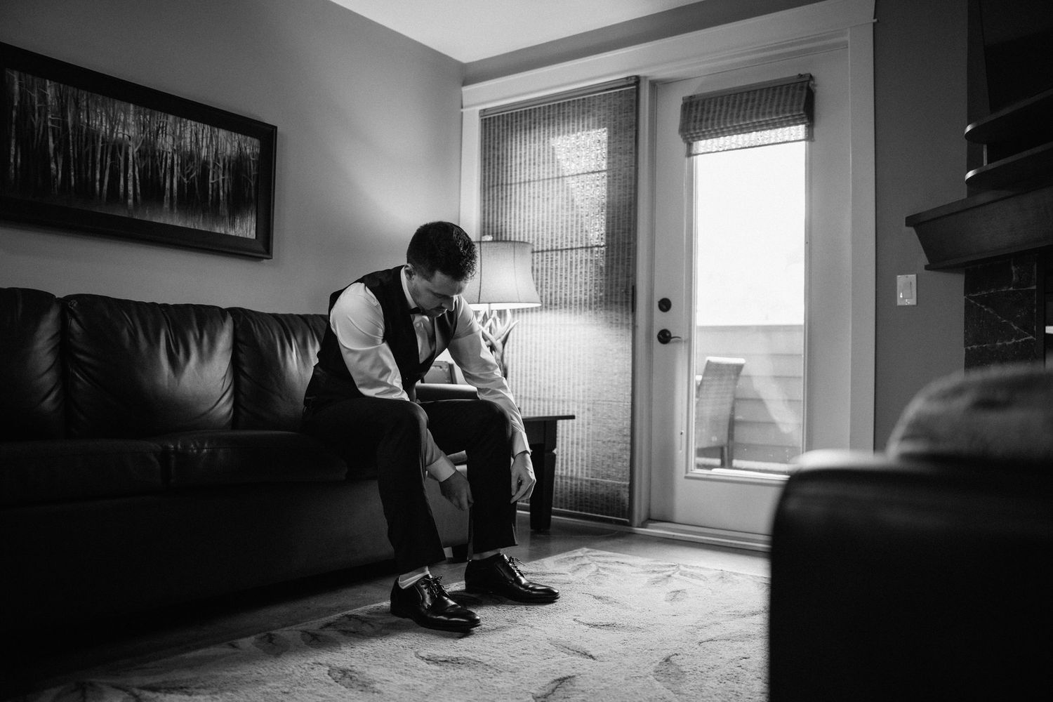 Black and white photo of the groom in a vest and bow tie putting on his shoes on a leather couch