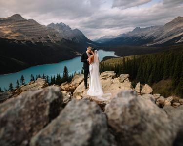 A couple shares a romantic kiss in wedding attire atop rocky cliffs overlooking a turquoise mountain lake.