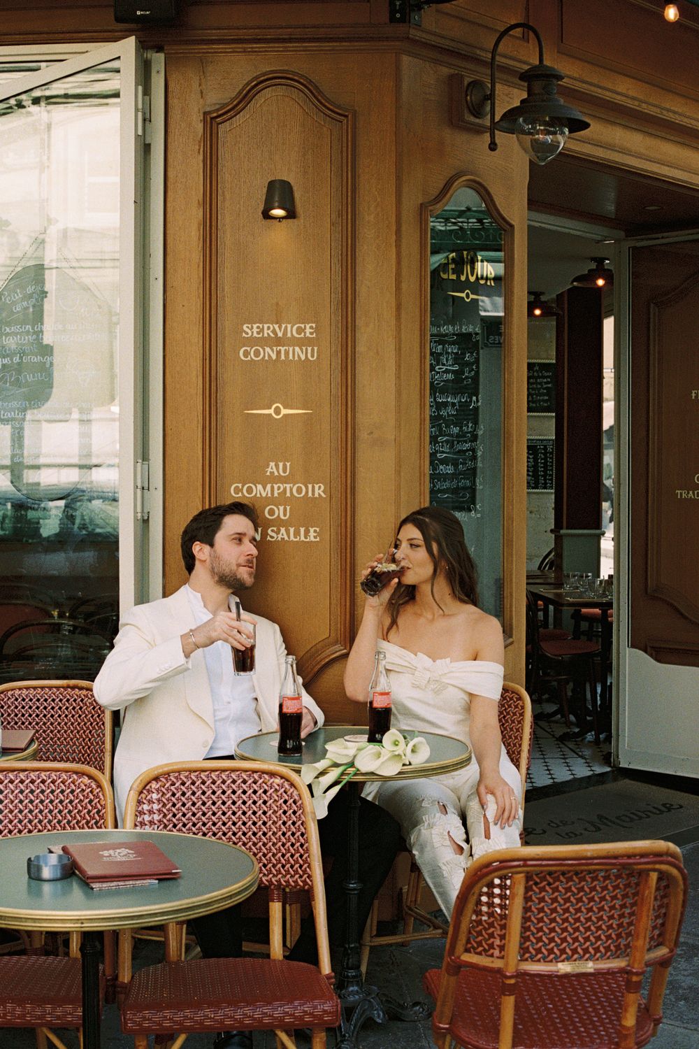 Elegant couple enjoying drinks at a charming Parisian cafe terrace with classic wicker chairs and wooden facade. Paris film wedding