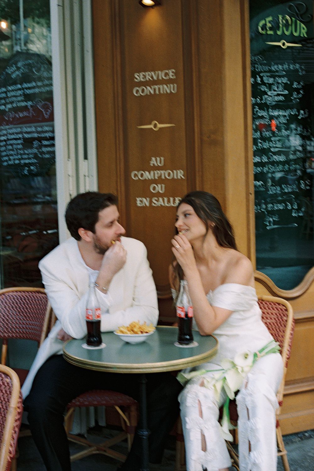A bride and groom share a romantic moment at a Parisian café terrace, enjoying Coca-Cola and fries together. Paris film wedding photos