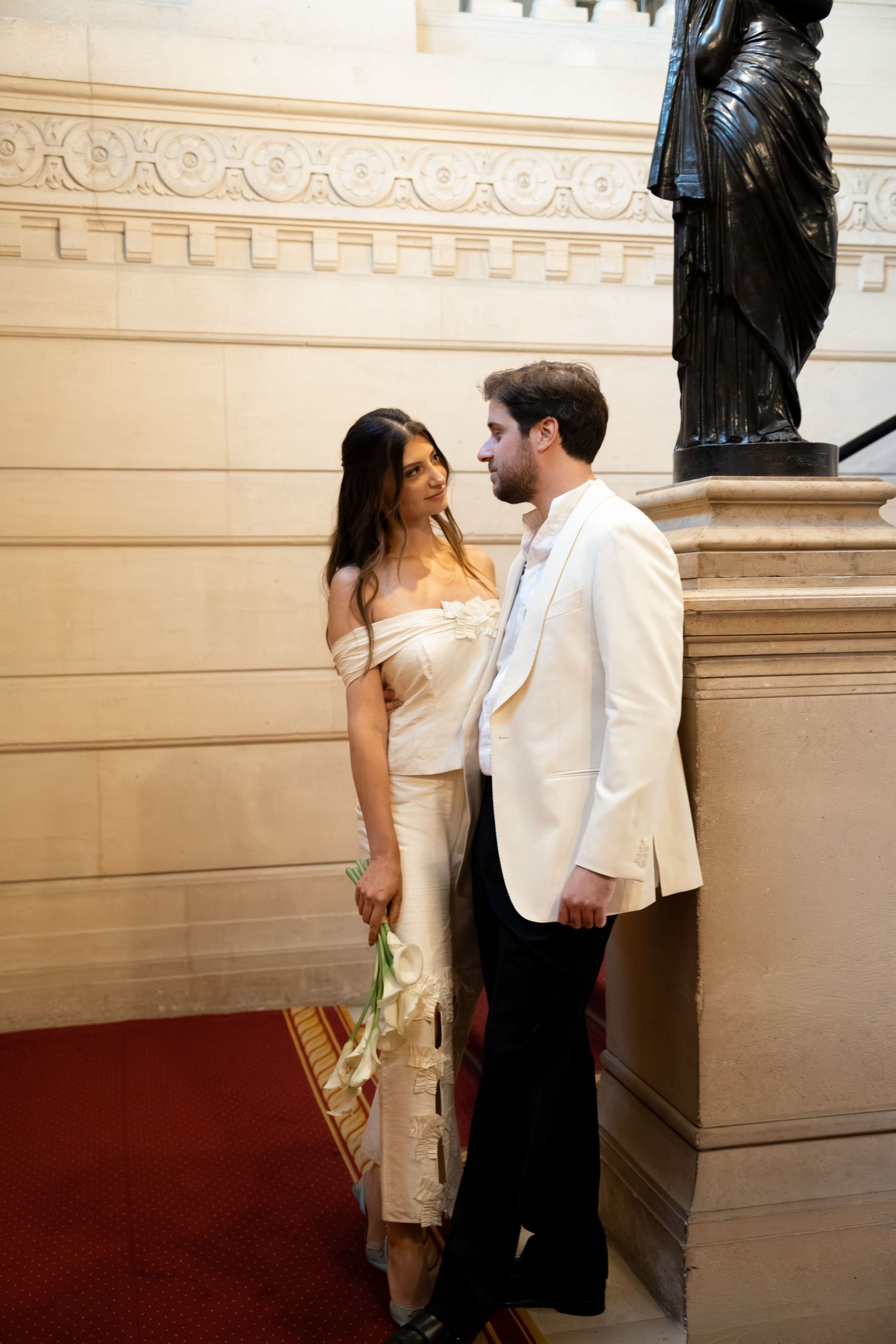 Elegant couple in formal attire standing near a bronze statue in a grand marble hall, woman holding white flowers.