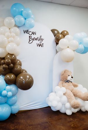 Baby shower balloon arch in blue, white, and brown with teddy bear and "We Can Bearly Wait" sign.