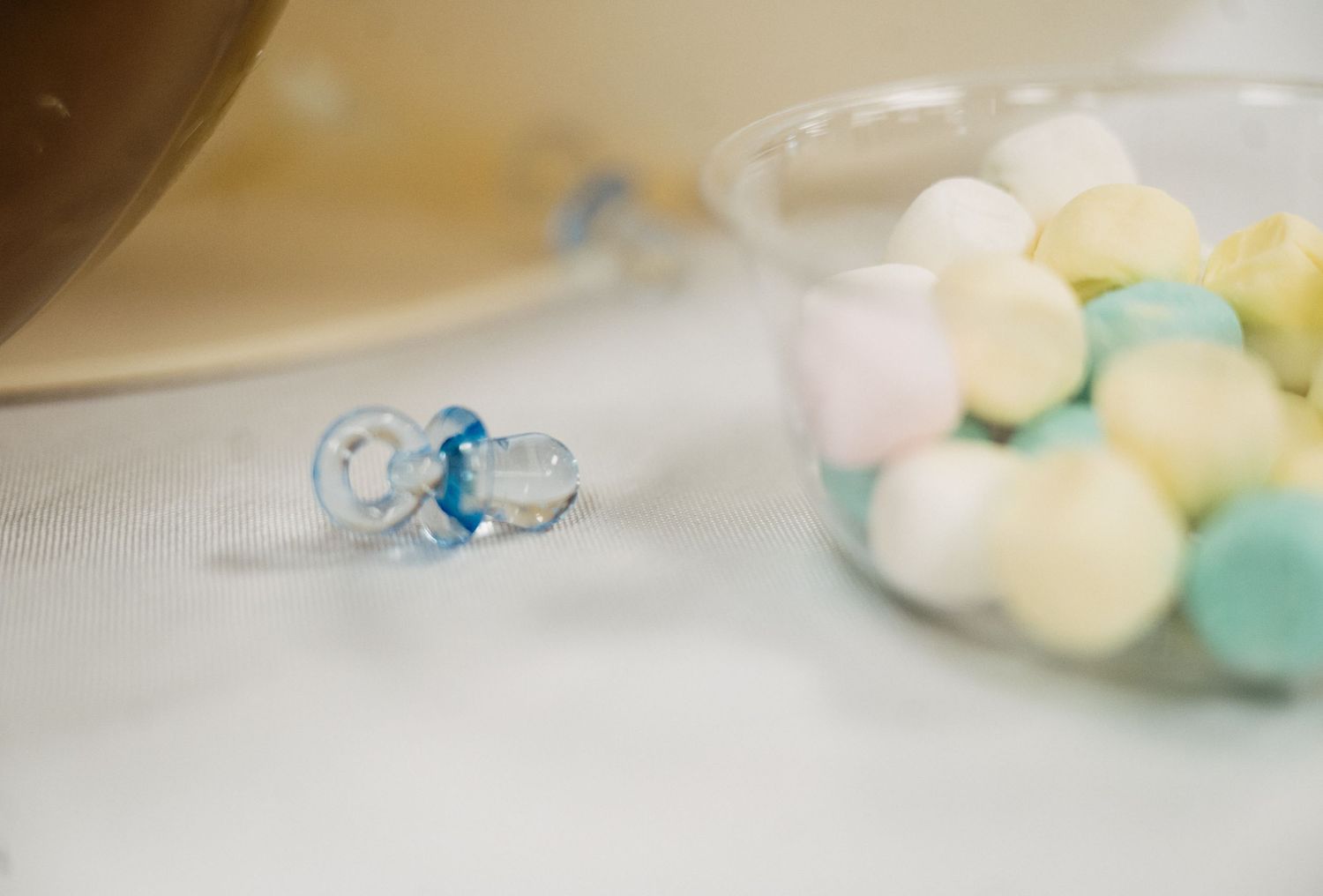 Blue baby pacifier next to a cup of colorful pastel candy balls on a white table at a baby shower.