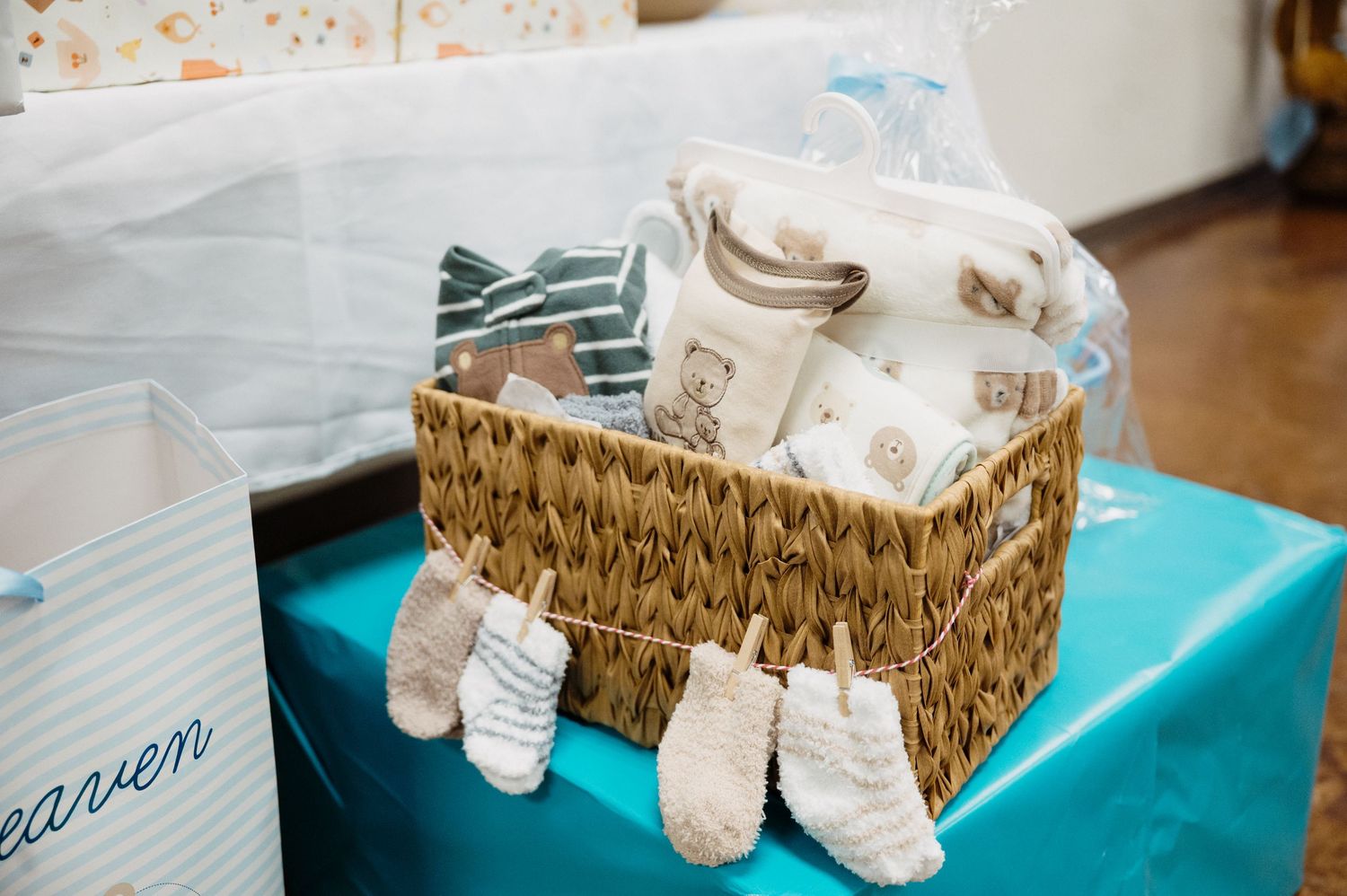 Wicker basket filled with baby shower gifts including clothing and accessories displayed on a teal table.