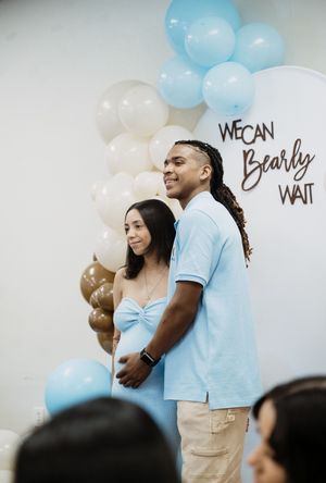 Couple at a baby shower with blue balloons and a 'We Can Bearly Wait' sign, expecting mother cradling her bump.