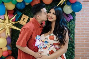 Couple posing at colorful balloon backdrop, man kissing pregnant woman's cheek, floral dress, festive gender reveal party.