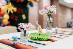 Colorful fiesta party table with rainbow runner, honeycomb centerpiece, dinosaur figurine, floral vases, and balloon decorations.