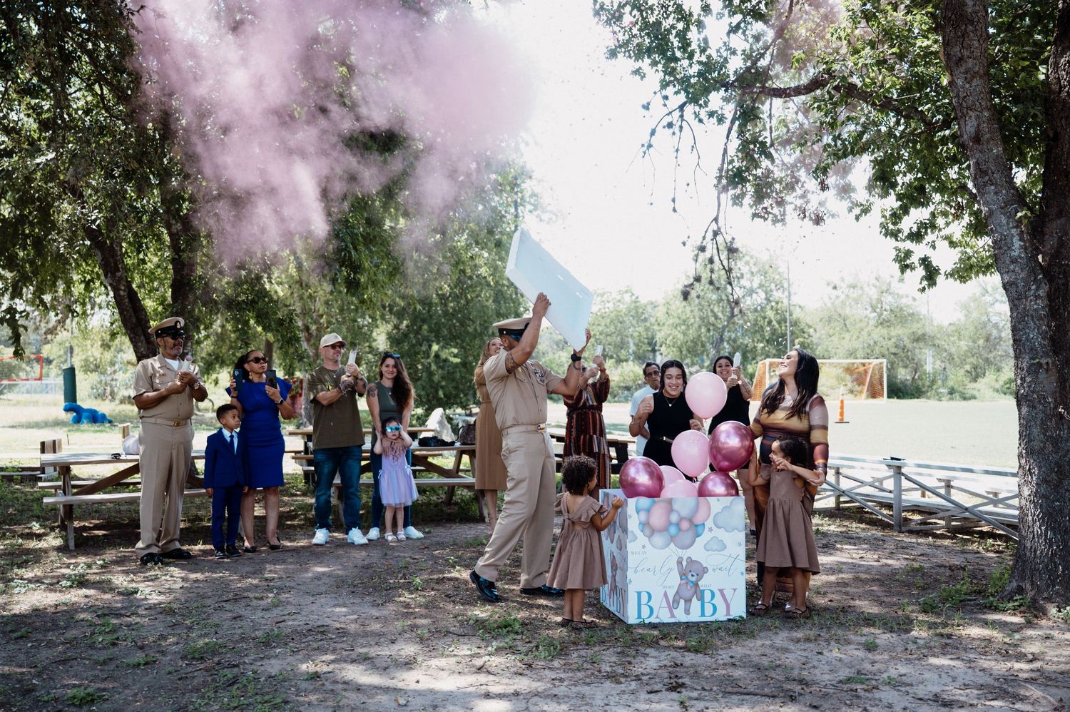 Gender reveal party with pink smoke cannon outdoors, guests gathered under trees celebrating a baby girl announcement.