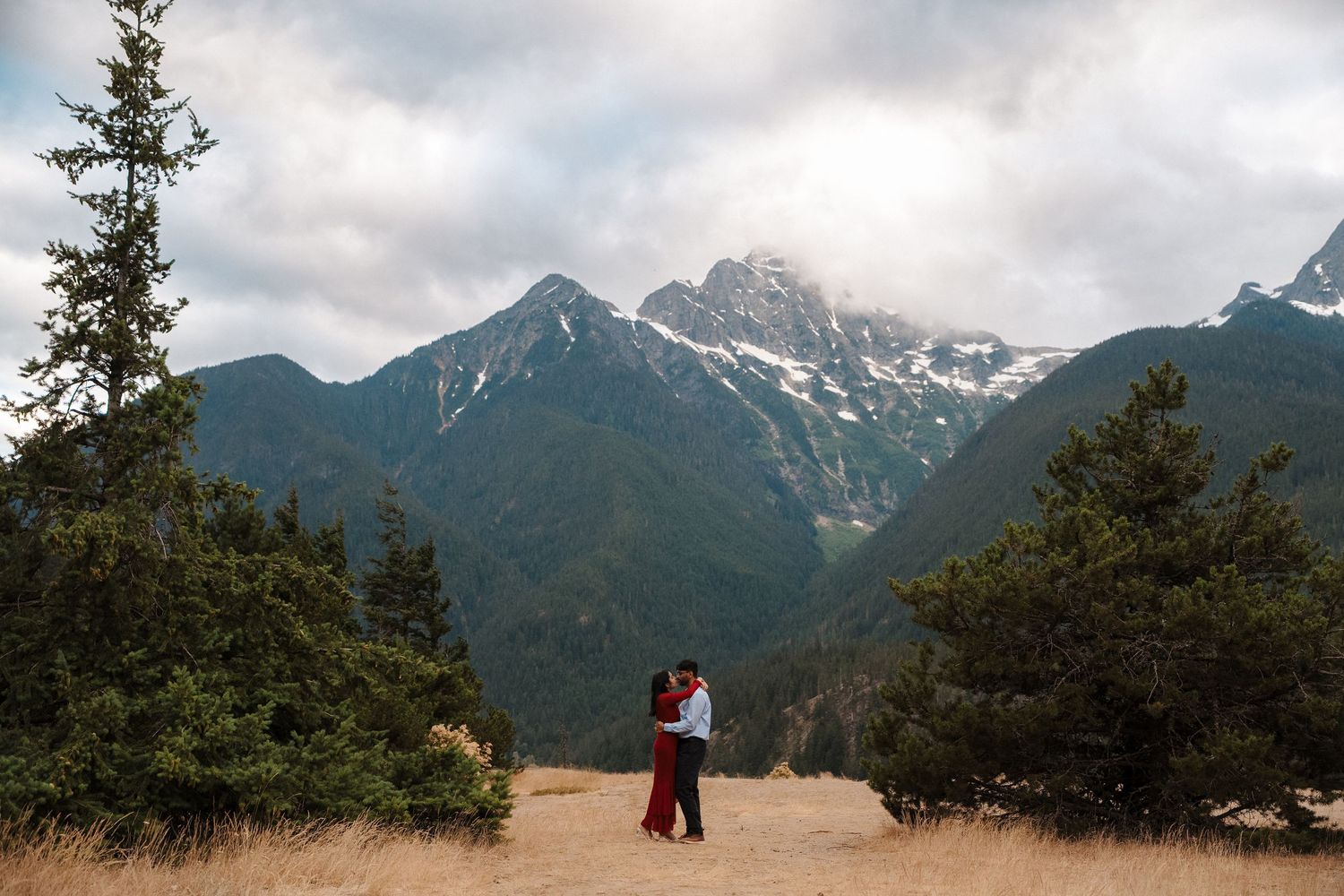 Couple embracing in mountain meadow with snow-capped peaks and evergreen trees under cloudy sky.