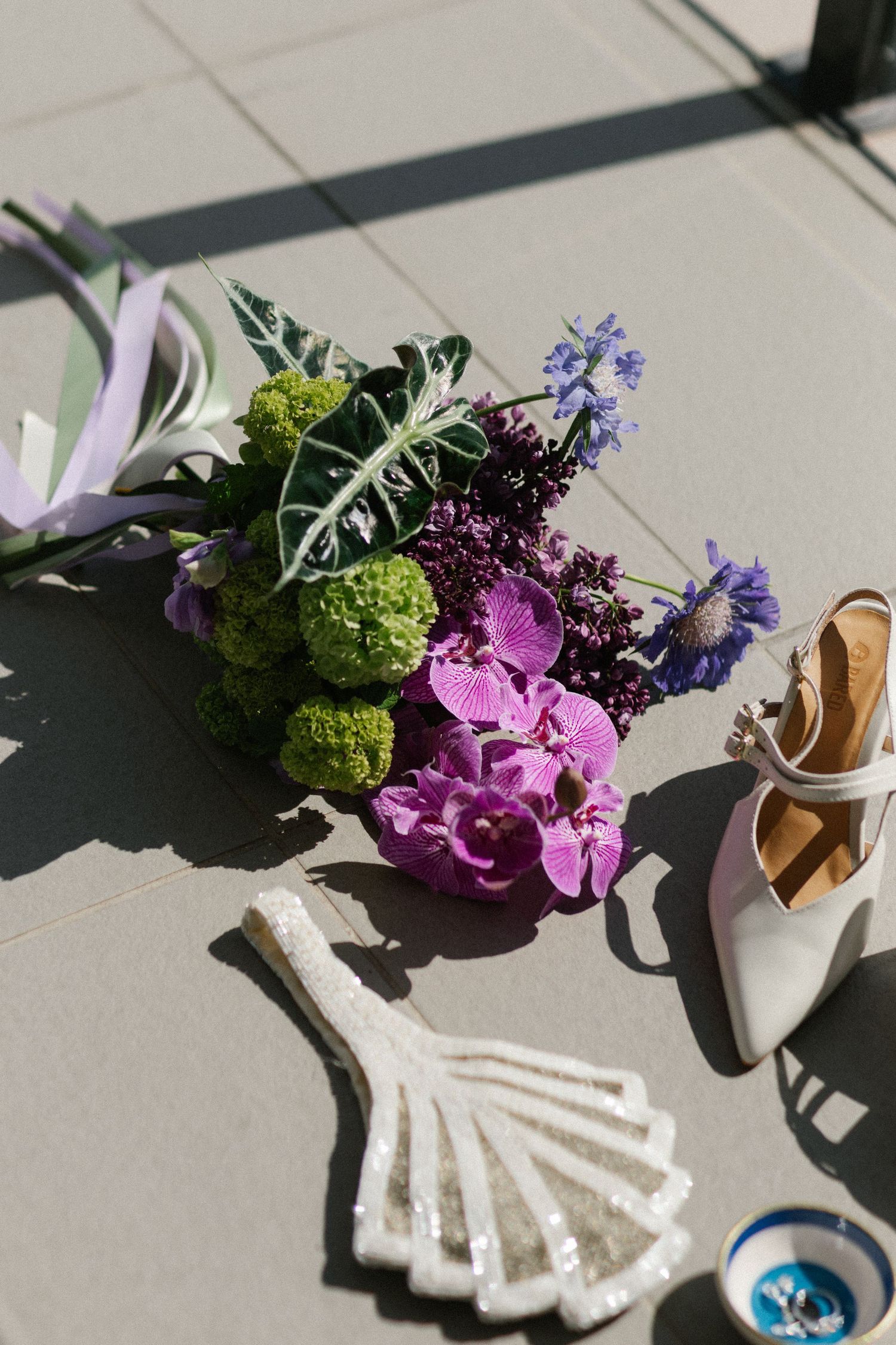 Bridal flat lay featuring purple orchid bouquet, white heels, and decorative fan on pavement.