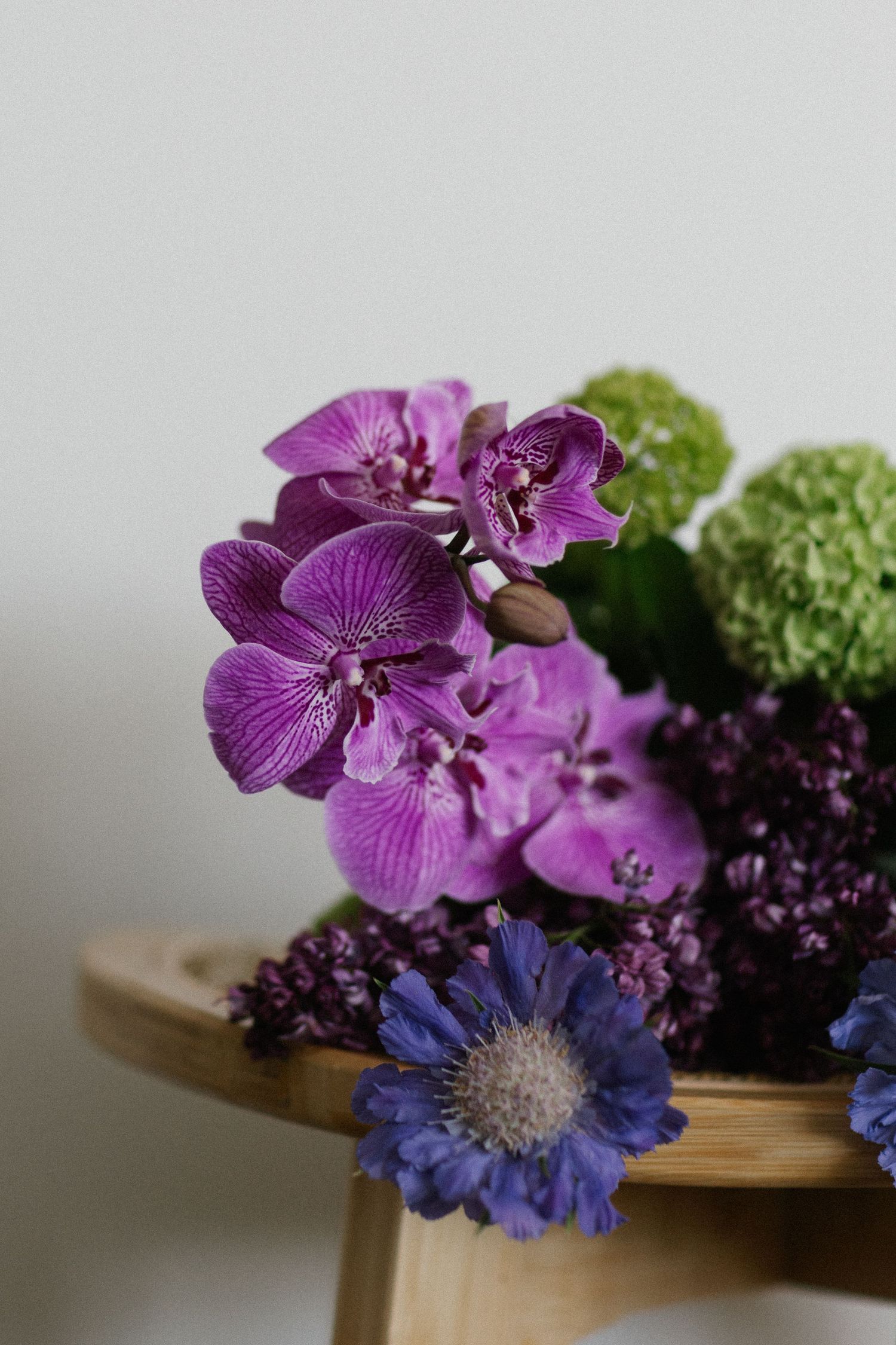 Elegant floral arrangement of purple orchids, lilac, and green hydrangeas on a wooden stand.