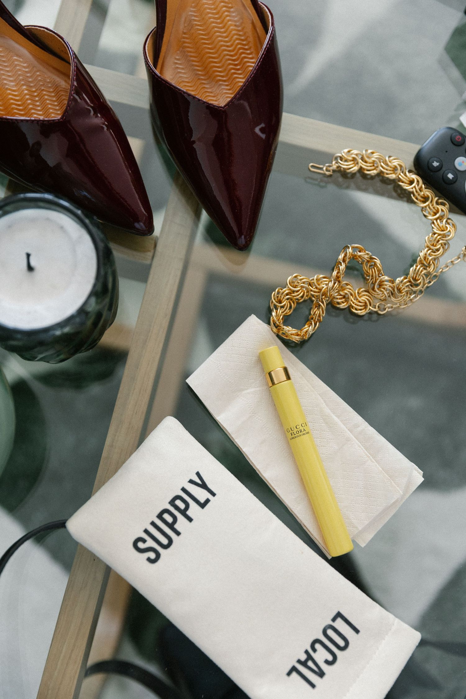 Flat lay of dark red patent heels, gold chain necklace, yellow perfume, and Supply Local pouch.