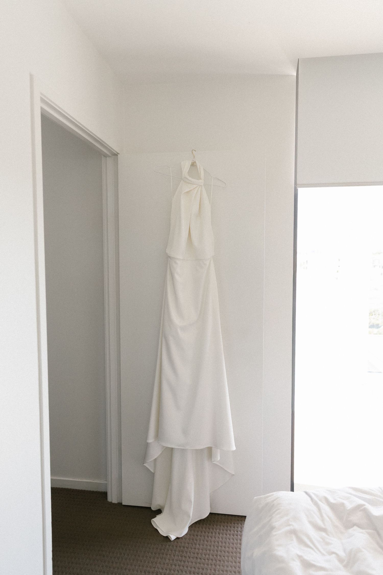 White halter neck wedding dress hanging on a hanger in a bright minimalist bedroom.