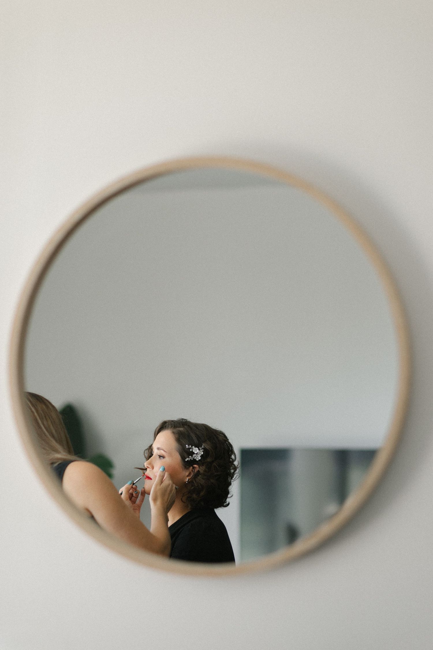 Bride getting lipstick applied by makeup artist reflected in a round wooden framed mirror.