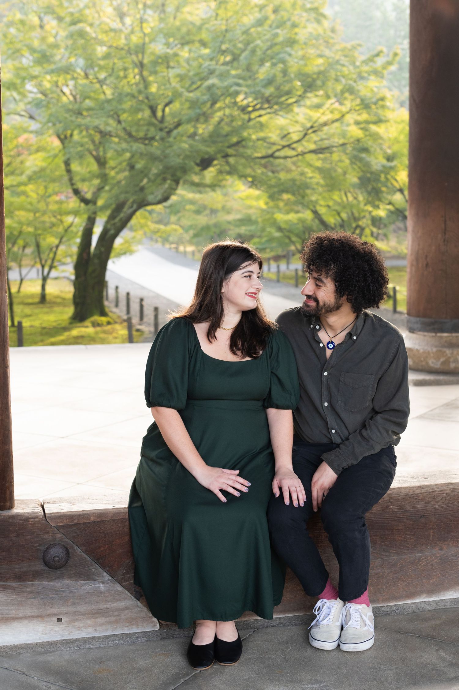 Couple sitting together on steps in a lush garden, woman in green dress, man in dark shirt, smiling at each other.
