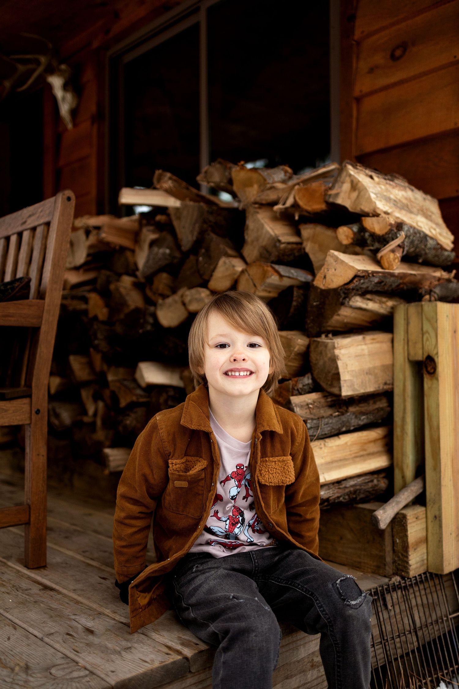 Smiling young boy in a rust-brown jacket and jeans sits on a wooden porch beside a neatly stacked pile of firewood at a rustic log cabin.