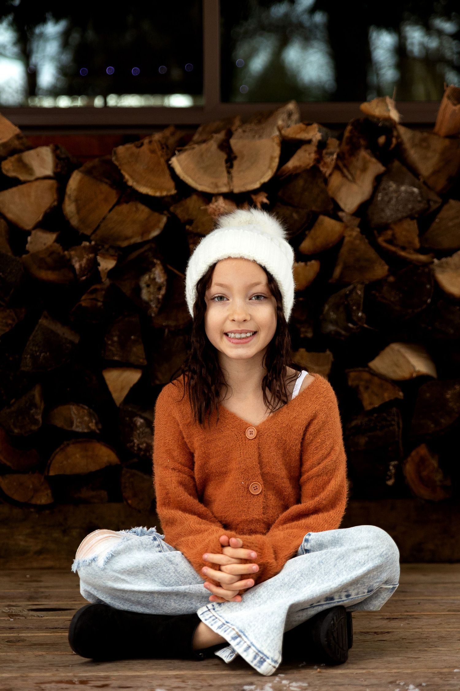 Smiling child in a white fuzzy hat and rust cardigan sits cross-legged on a wooden deck in front of a stacked firewood pile.