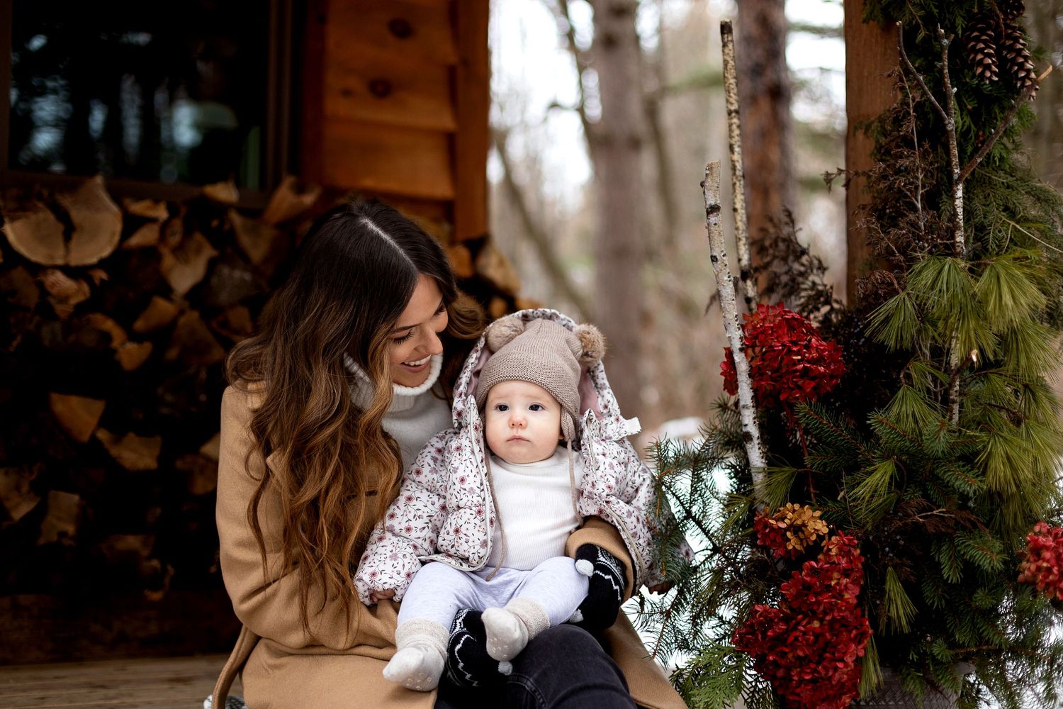 Smiling woman in a tan coat holds a baby in a knit hat and snowsuit beside a Christmas tree with red berries near a stacked firewood cabin.