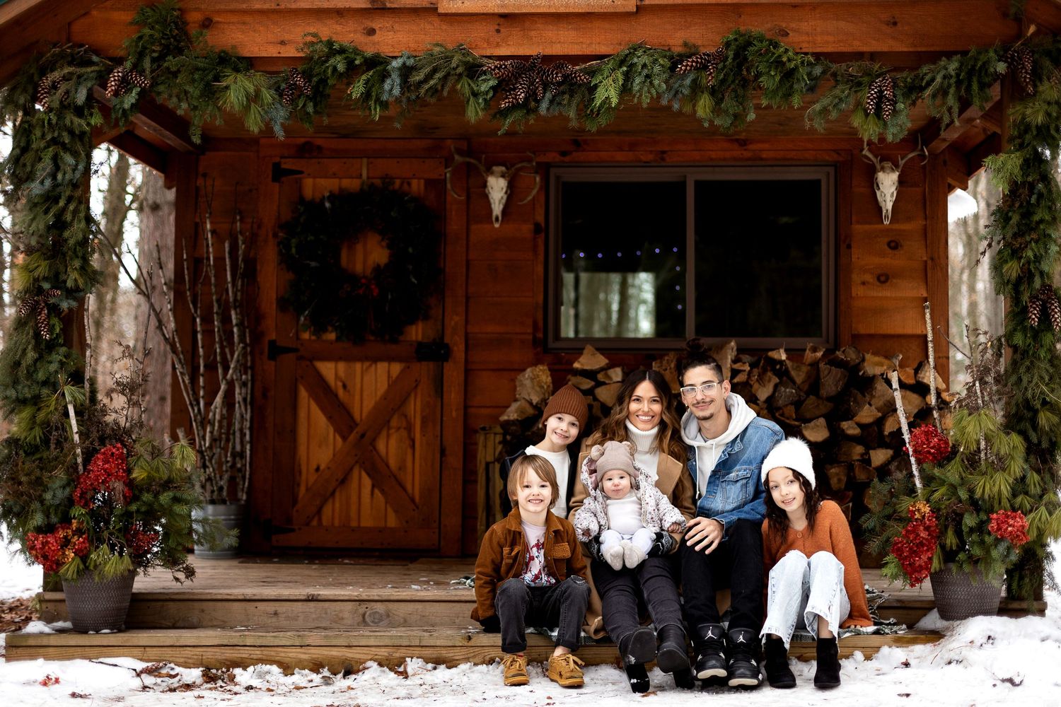 Family of six sits together on the porch of a rustic log cabin decorated with Christmas garlands, wreaths and poinsettias in the snow.