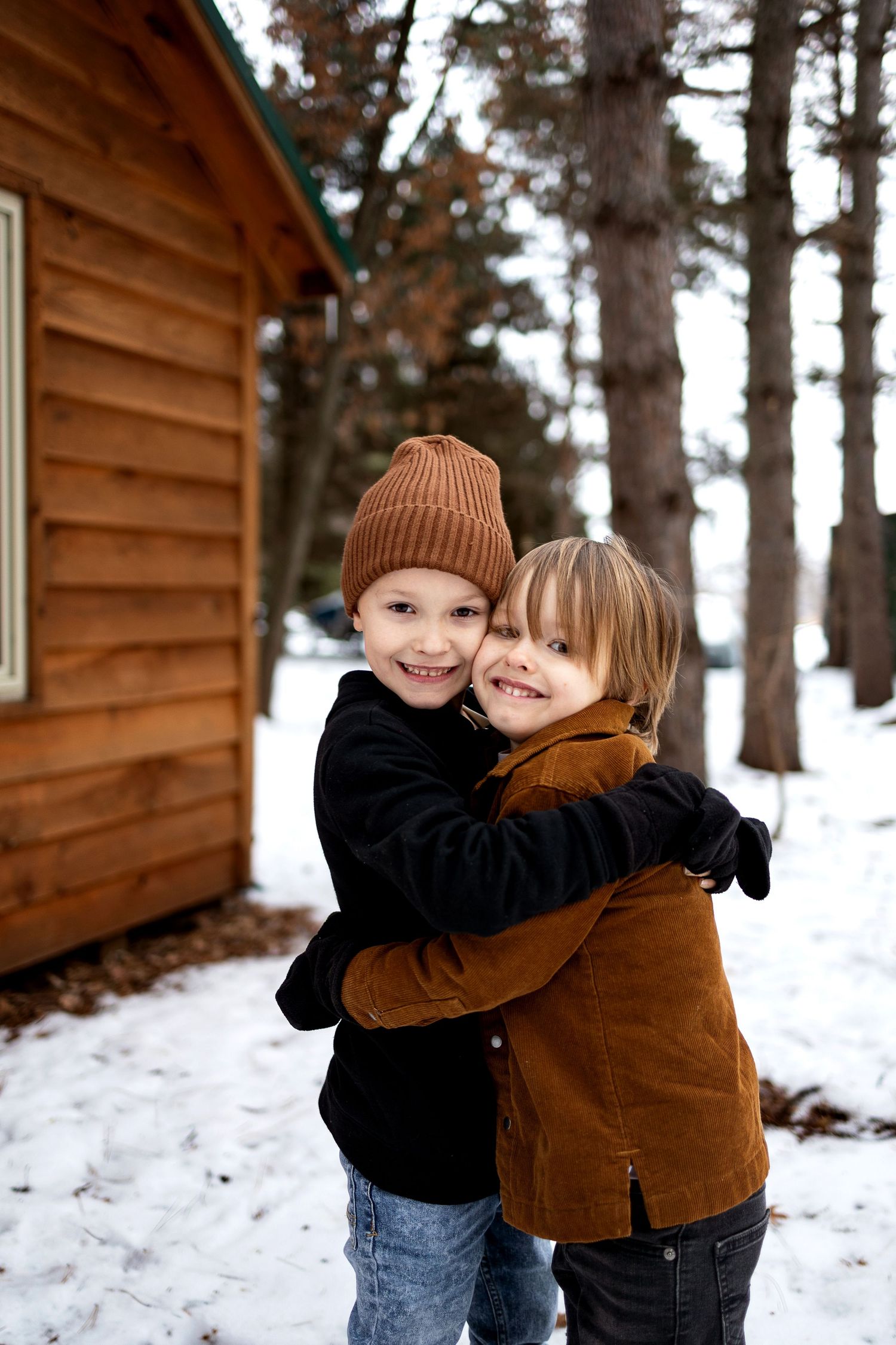 Two smiling children hug outdoors in the snow beside a log cabin, one in a brown knit beanie and one in a rust corduroy jacket.