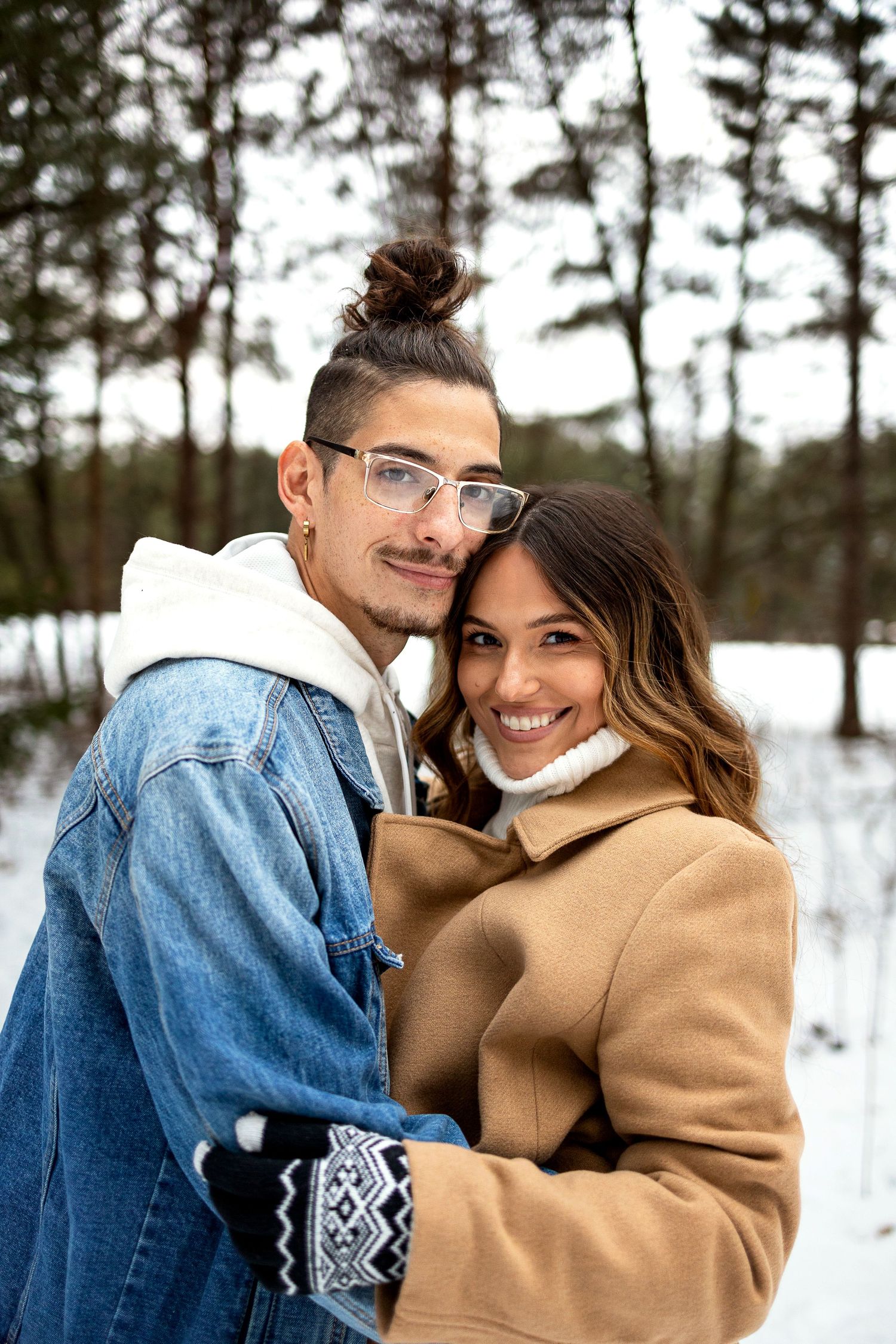 Smiling couple embracing in a snowy pine forest, he wears a denim jacket and glasses, she wears a tan coat and white scarf.