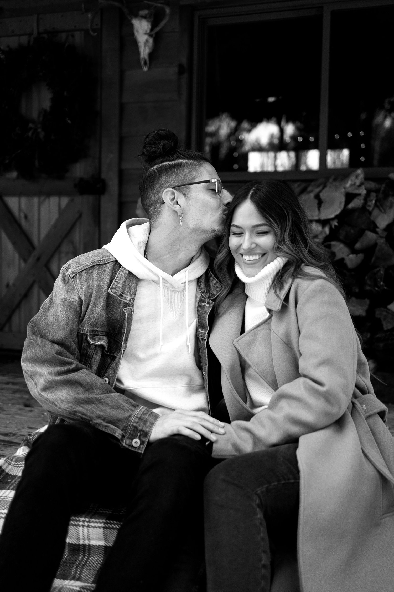 Black and white photo of a man in a denim jacket and hoodie kissing a smiling woman in a long coat on a rustic porch.