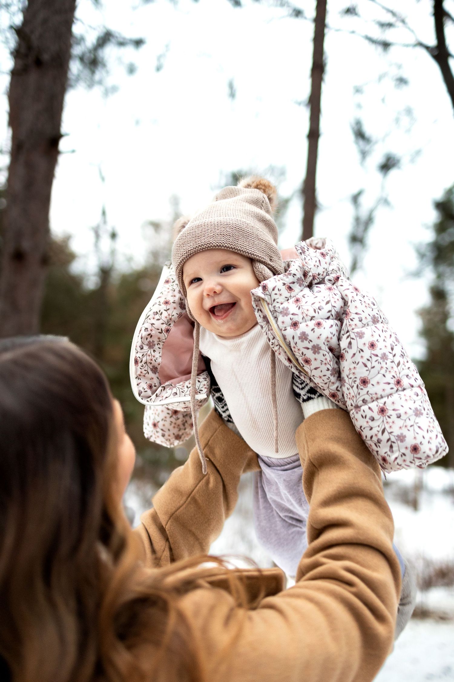 Smiling baby in a knit hat and floral jacket is lifted high by a mother in a camel coat among snowy winter trees.