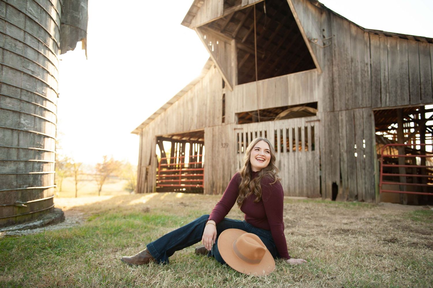 High school senior in burgundy top and jeans sits on grass before a rustic wooden barn at golden hour at Harlinsdale Farm