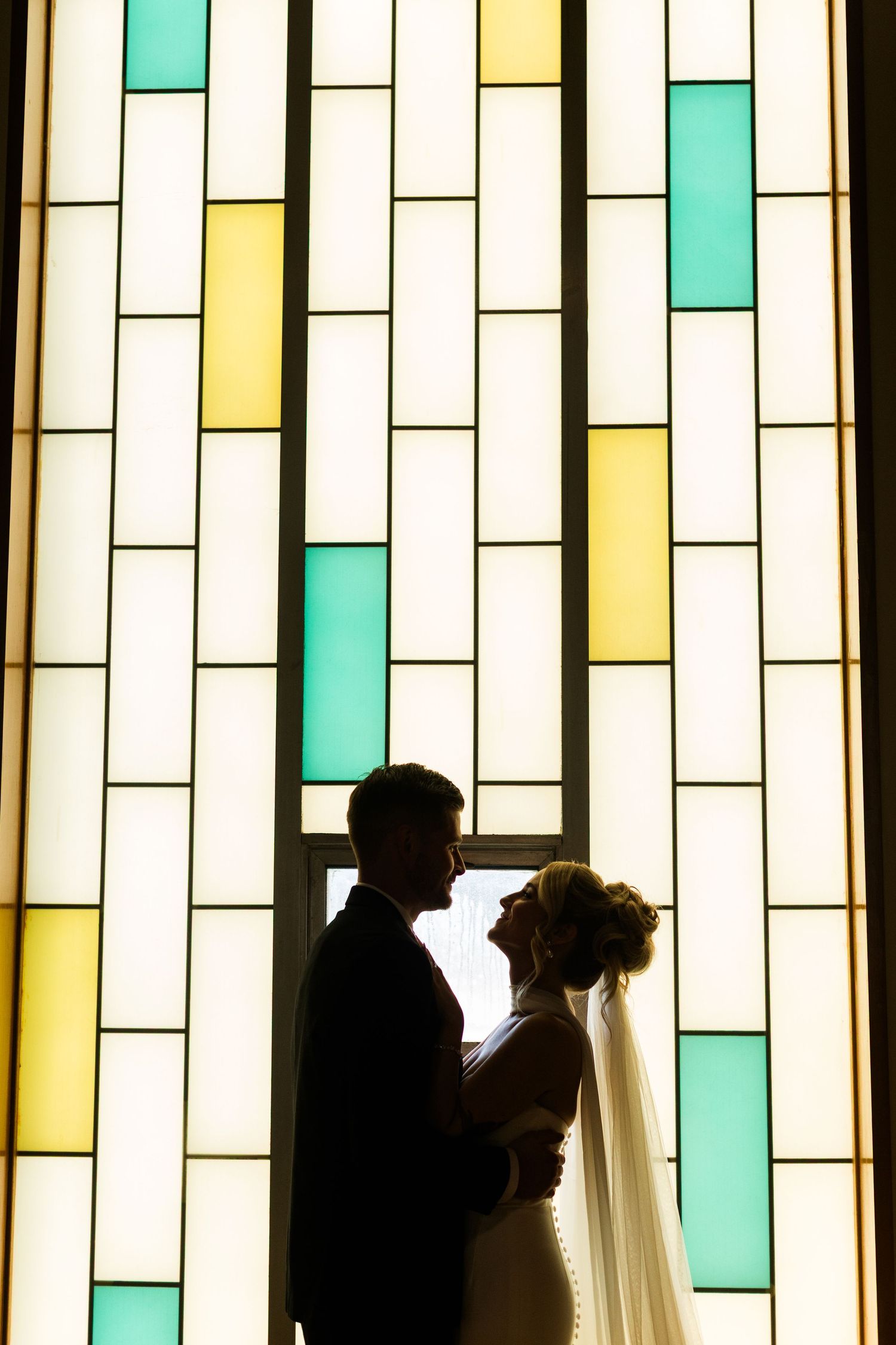 Silhouette of bride and groom embracing before a colorful stained glass window in teal, yellow, and white hues.