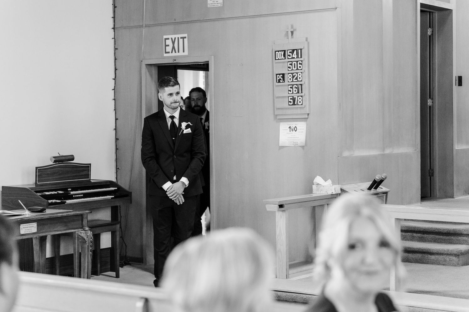 Groom stands at church entrance in black tuxedo, awaiting wedding ceremony in black and white photo.