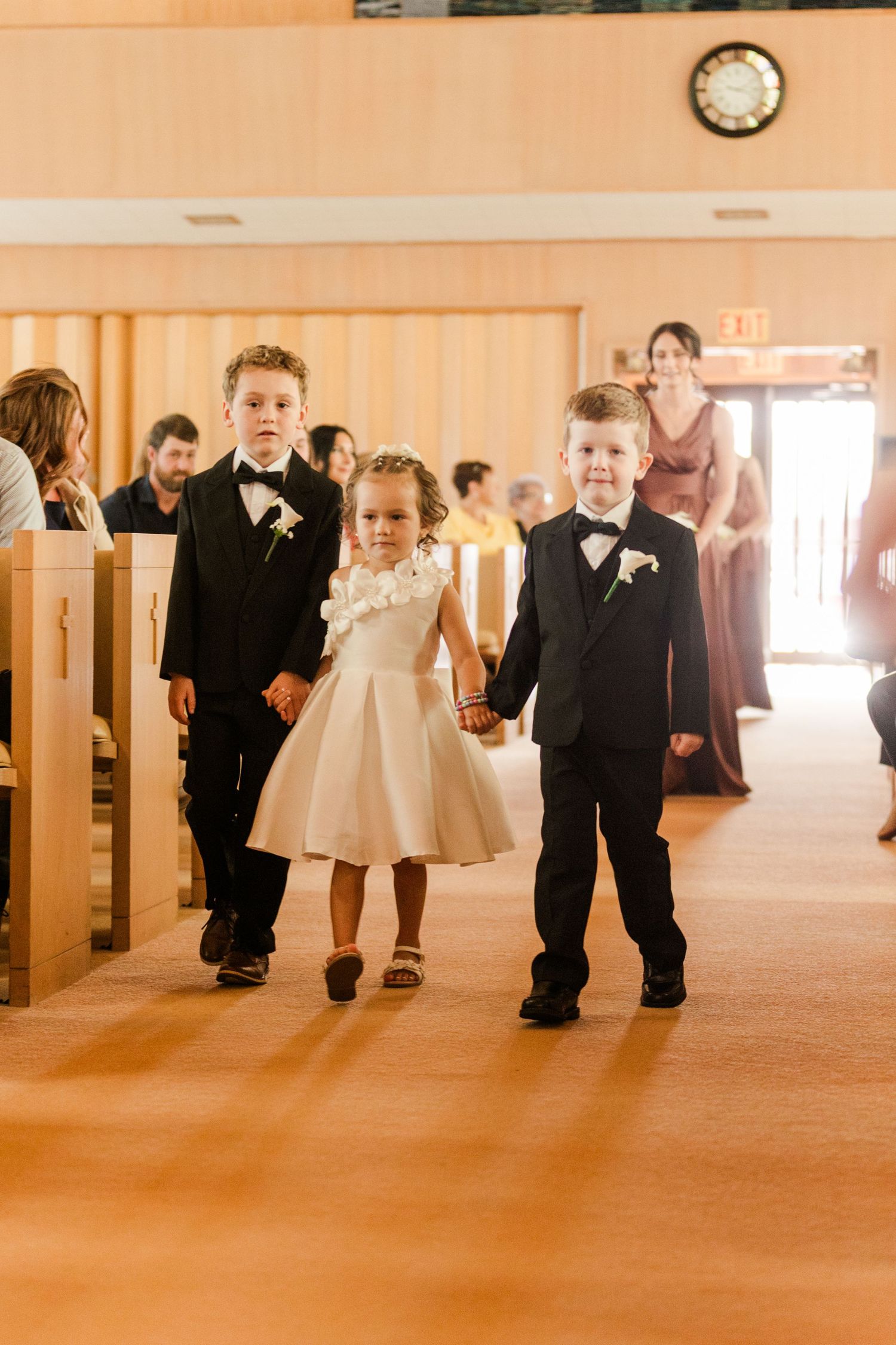Two ring bearers in tuxedos and a flower girl in white dress walk down church aisle during wedding ceremony.