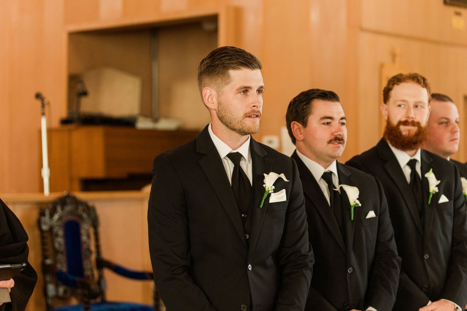 Groomsmen in black suits with white boutonnieres stand at the altar during a wedding ceremony.