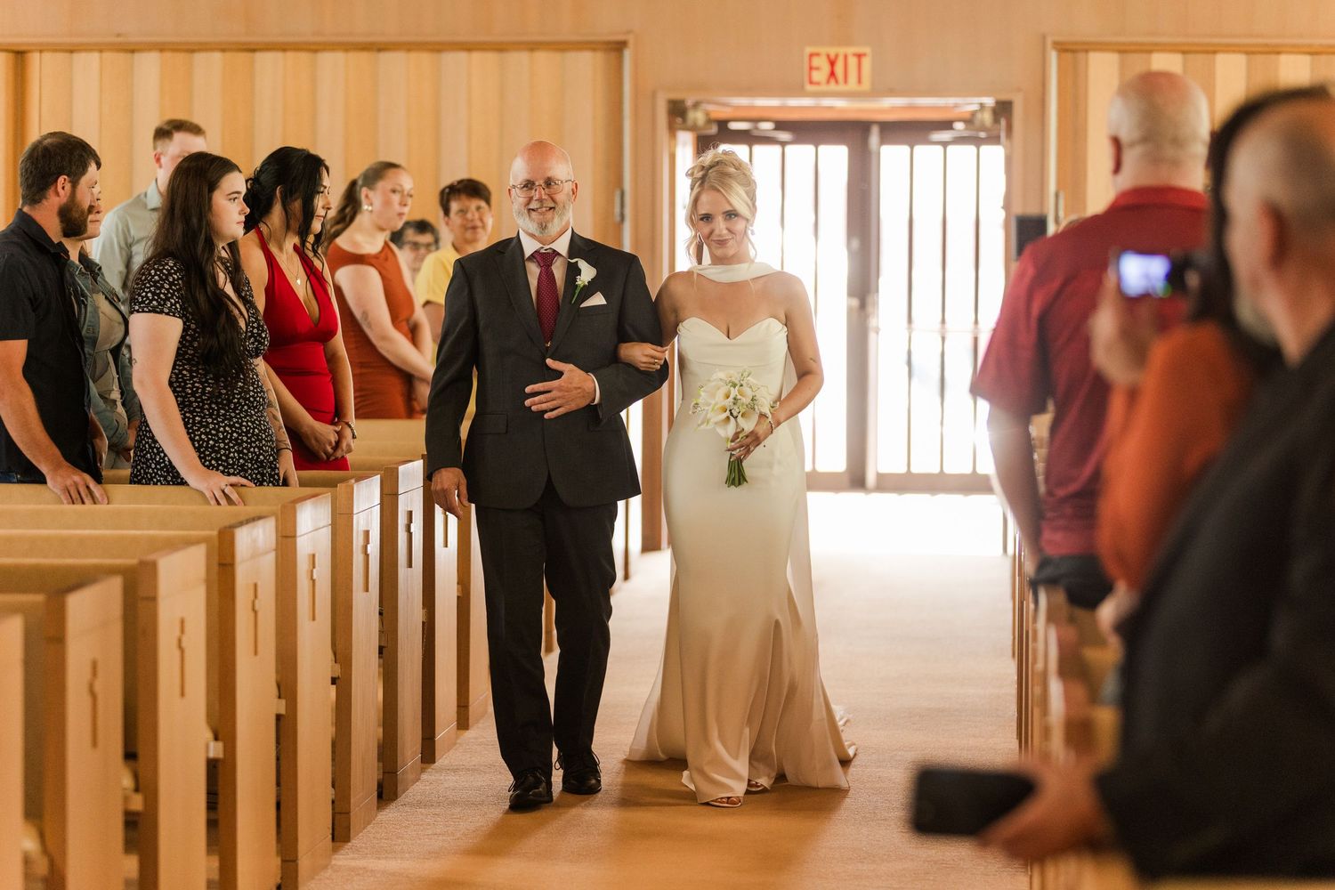 Bride walks down the aisle with her father in a wooden chapel, guests in red attire lining the pews.