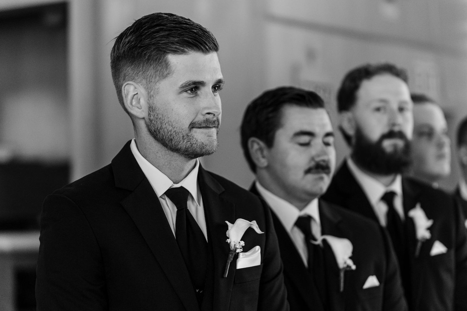 Black and white photo of groomsmen in suits with boutonnieres, groom in foreground looking emotional at wedding ceremony.