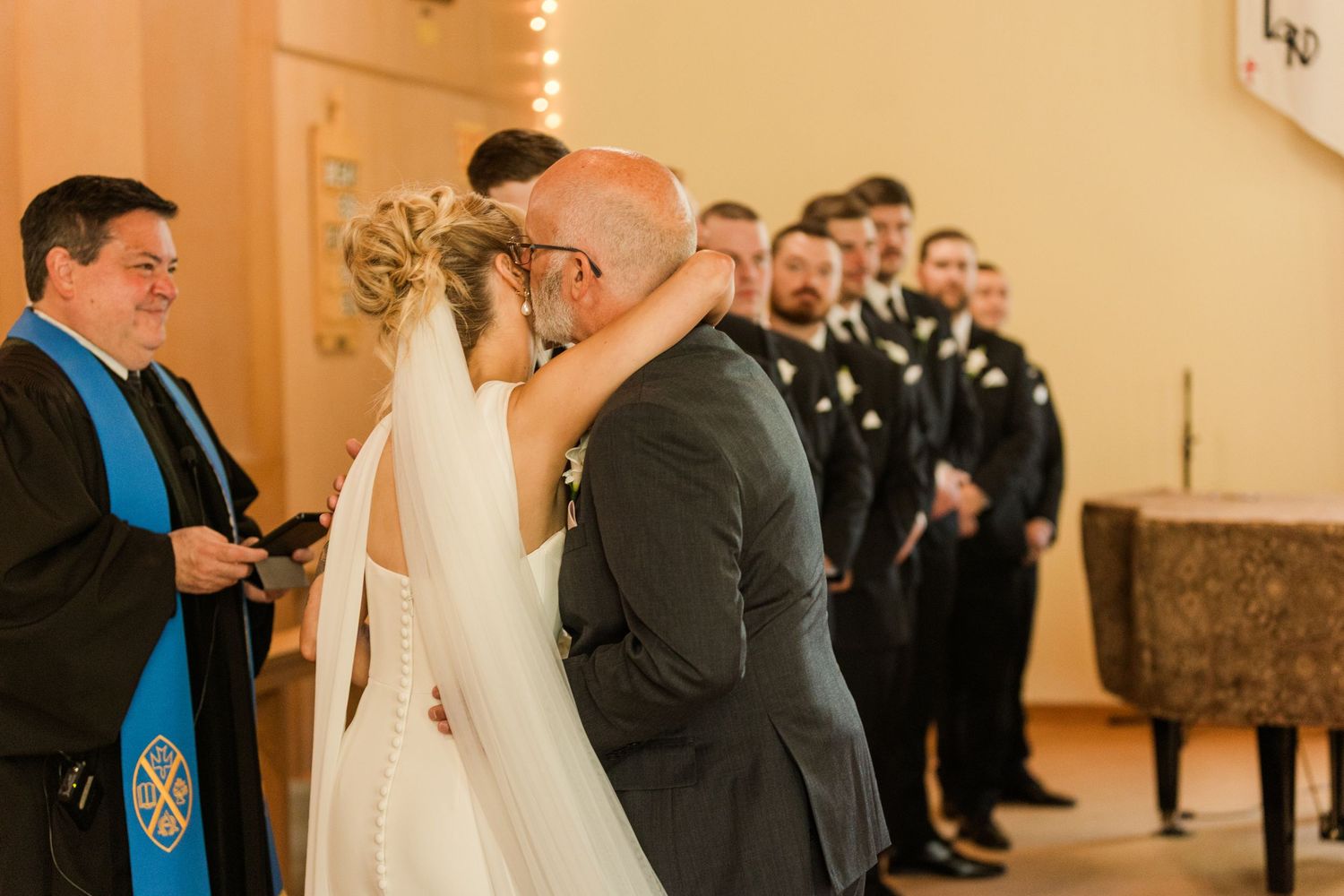 Bride and groom share first kiss at wedding altar with officiant and groomsmen standing nearby in church ceremony.
