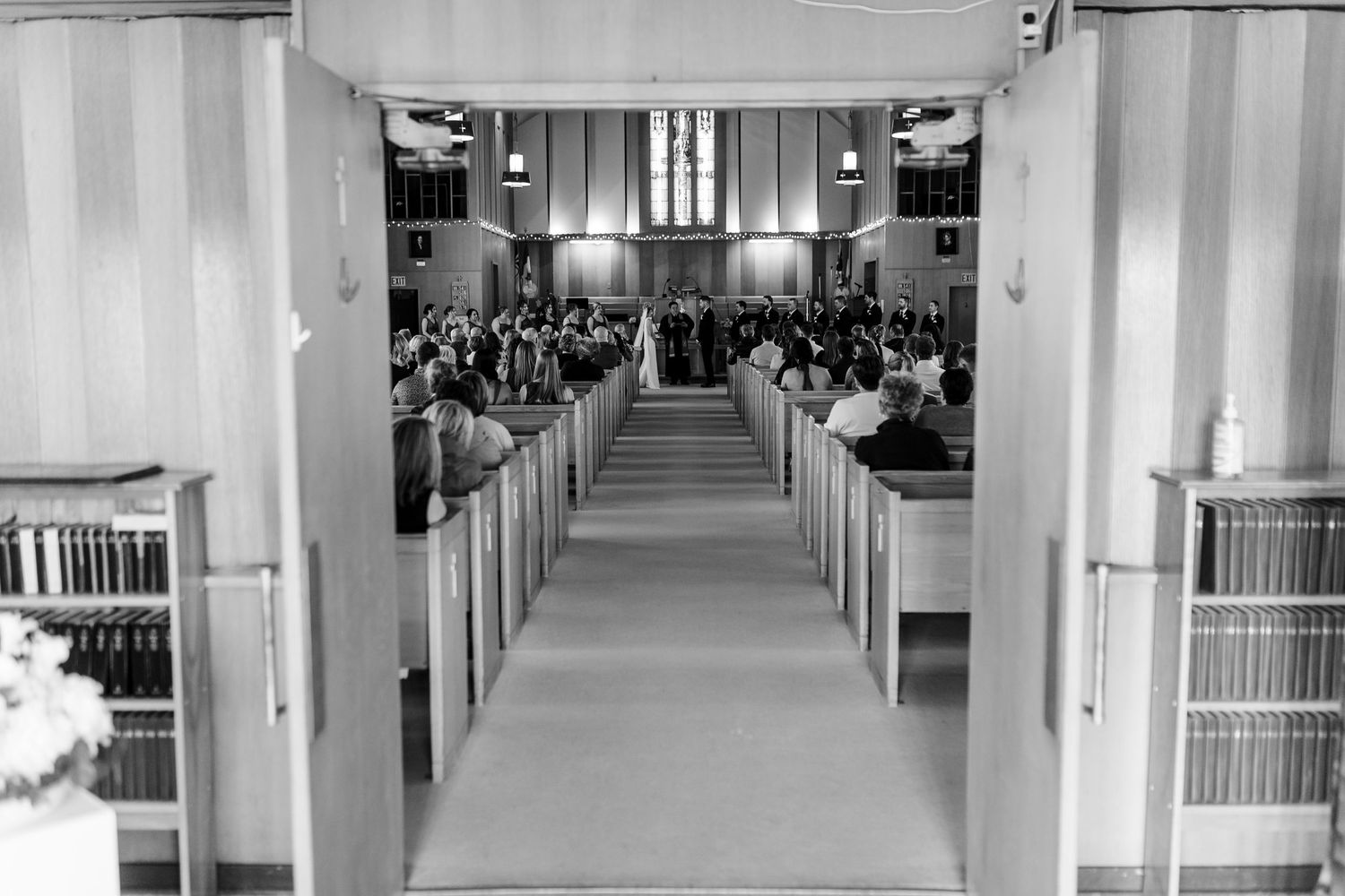 Black and white photo of a wedding ceremony viewed from the back of a church aisle with guests seated in pews.